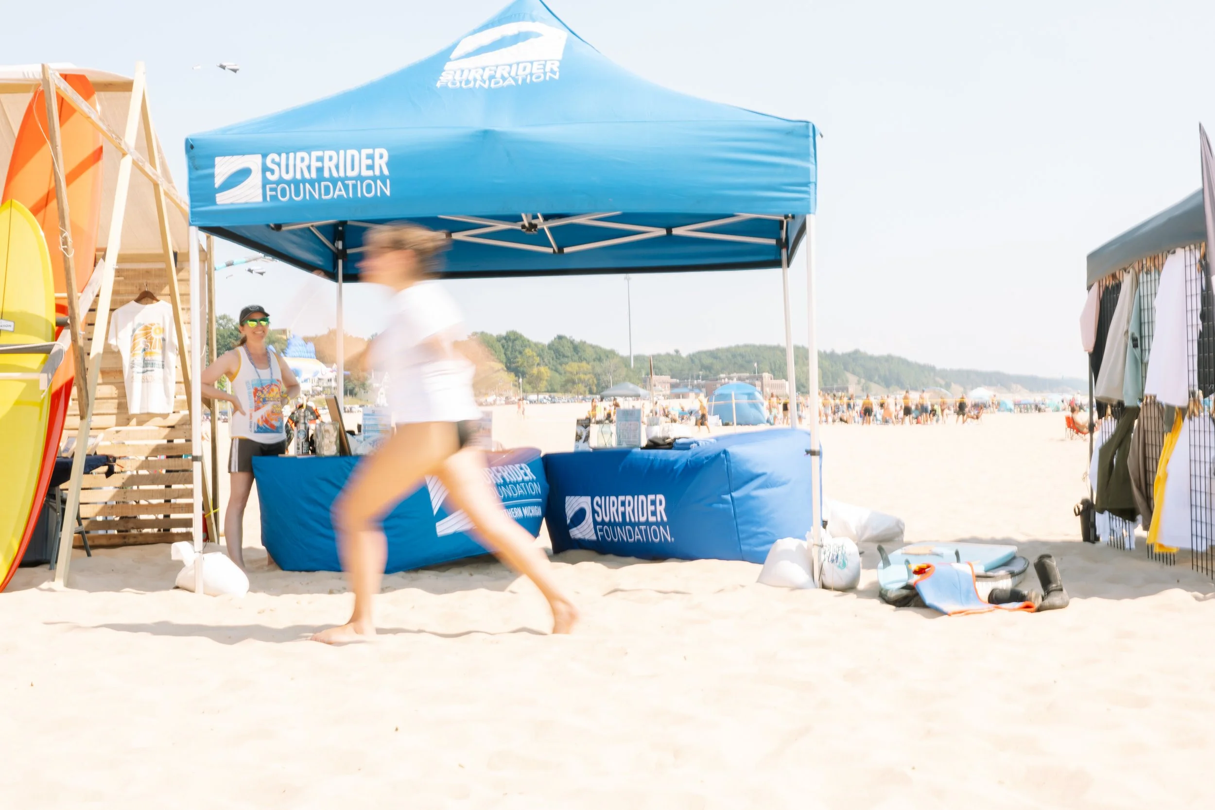 A beach scene with a blue tent and banner for the Surfrider Foundation. A woman in sunglasses stands near the tent, and a person in a white shirt is walking in front, blurred by motion. Other beachgoers and tents are in the background.