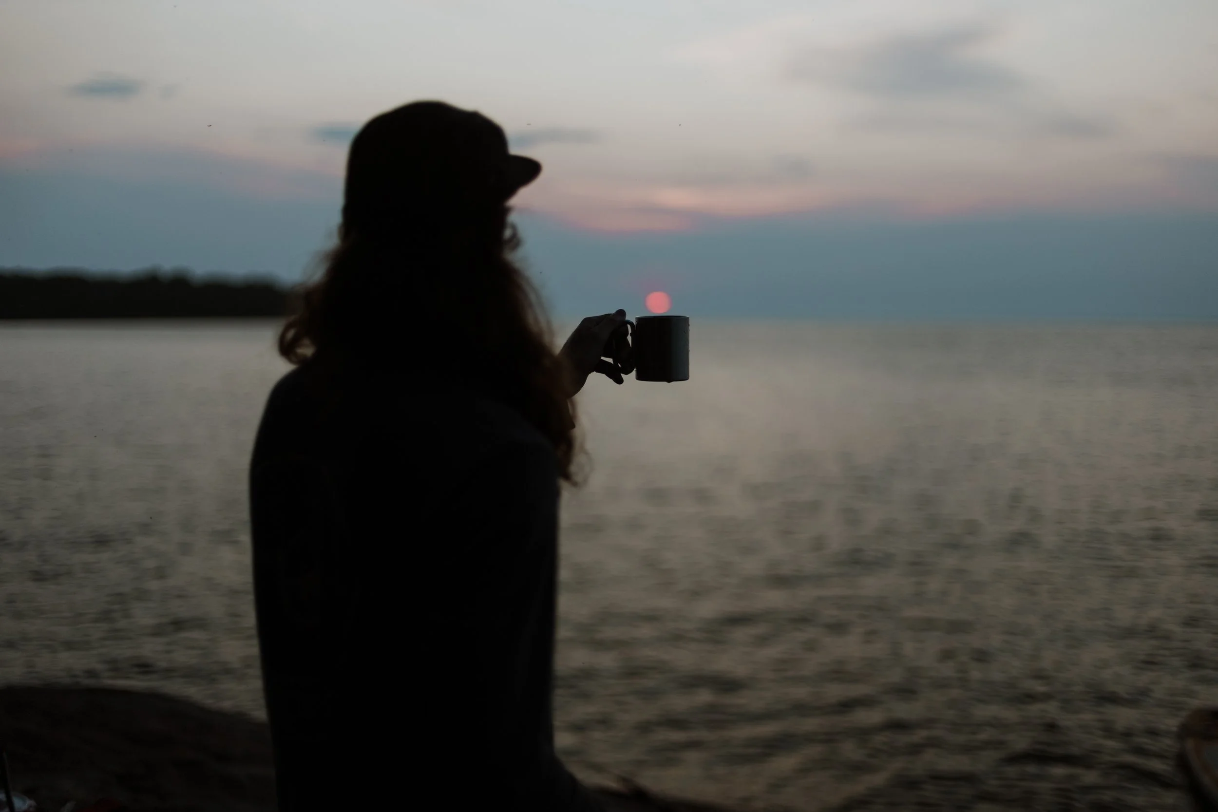 Silhouette of a person holding a mug, sitting by a body of water during sunset, with the sun near the horizon and a cloudy sky.