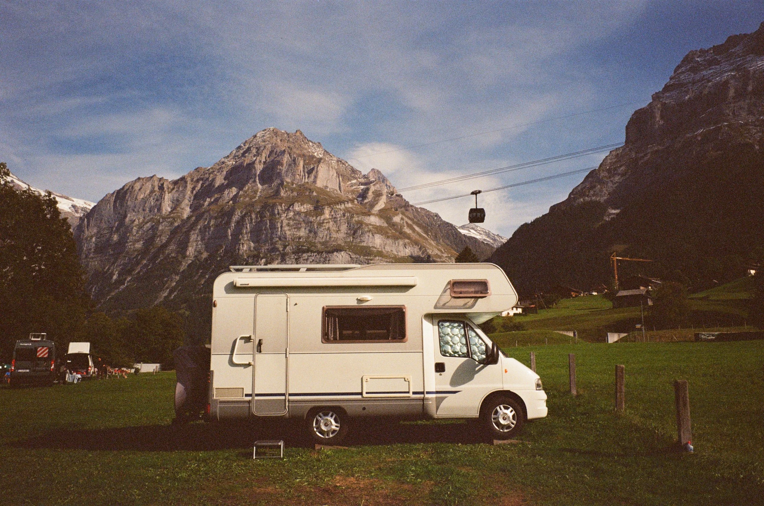 A white camper van parked on grass with mountains in the background and cable cars overhead.