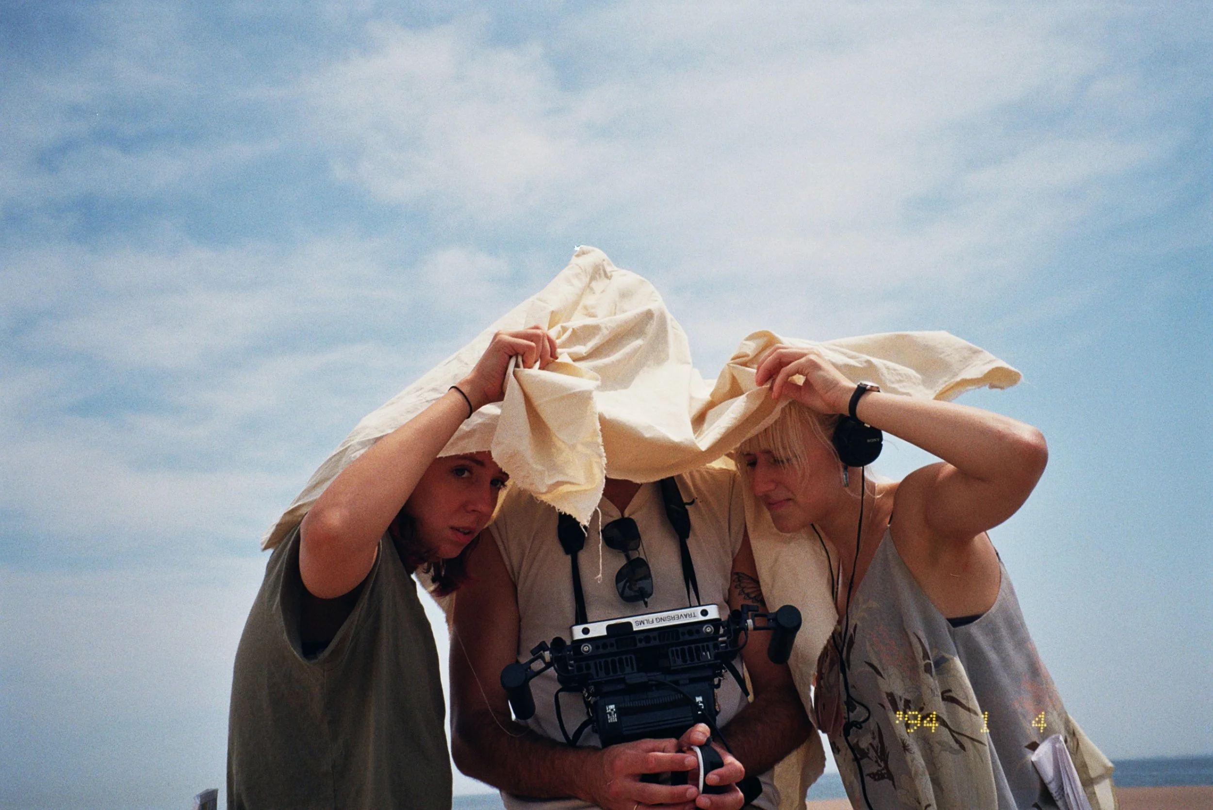 Three people, two women and one man, under a white cloth on a beach with a cloudy sky, with the man holding a camera, as they look at something under the cloth.