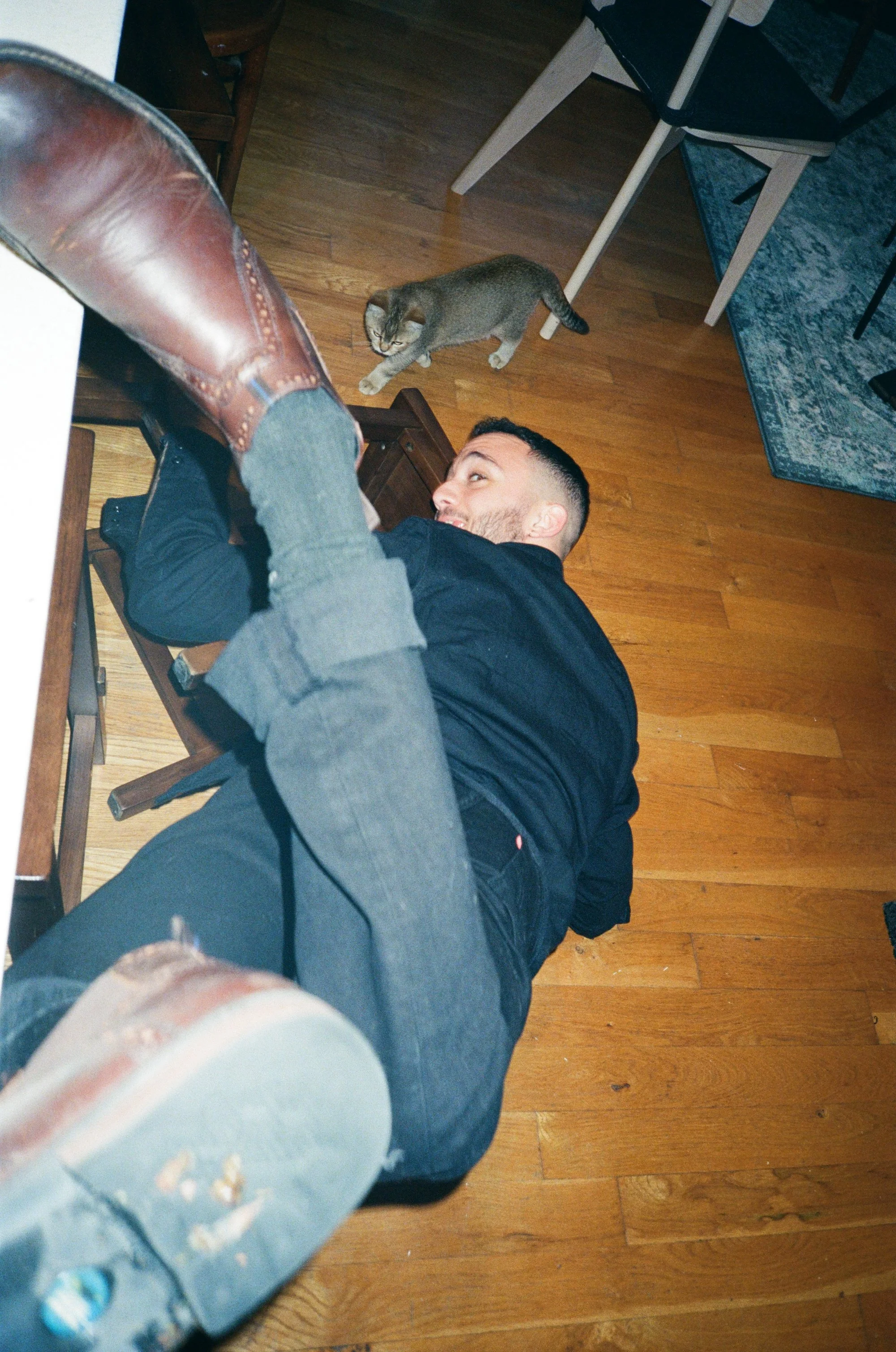 A man lying on the floor with a cat nearby, a person standing over him with a boot that is pressing on the man's face.
