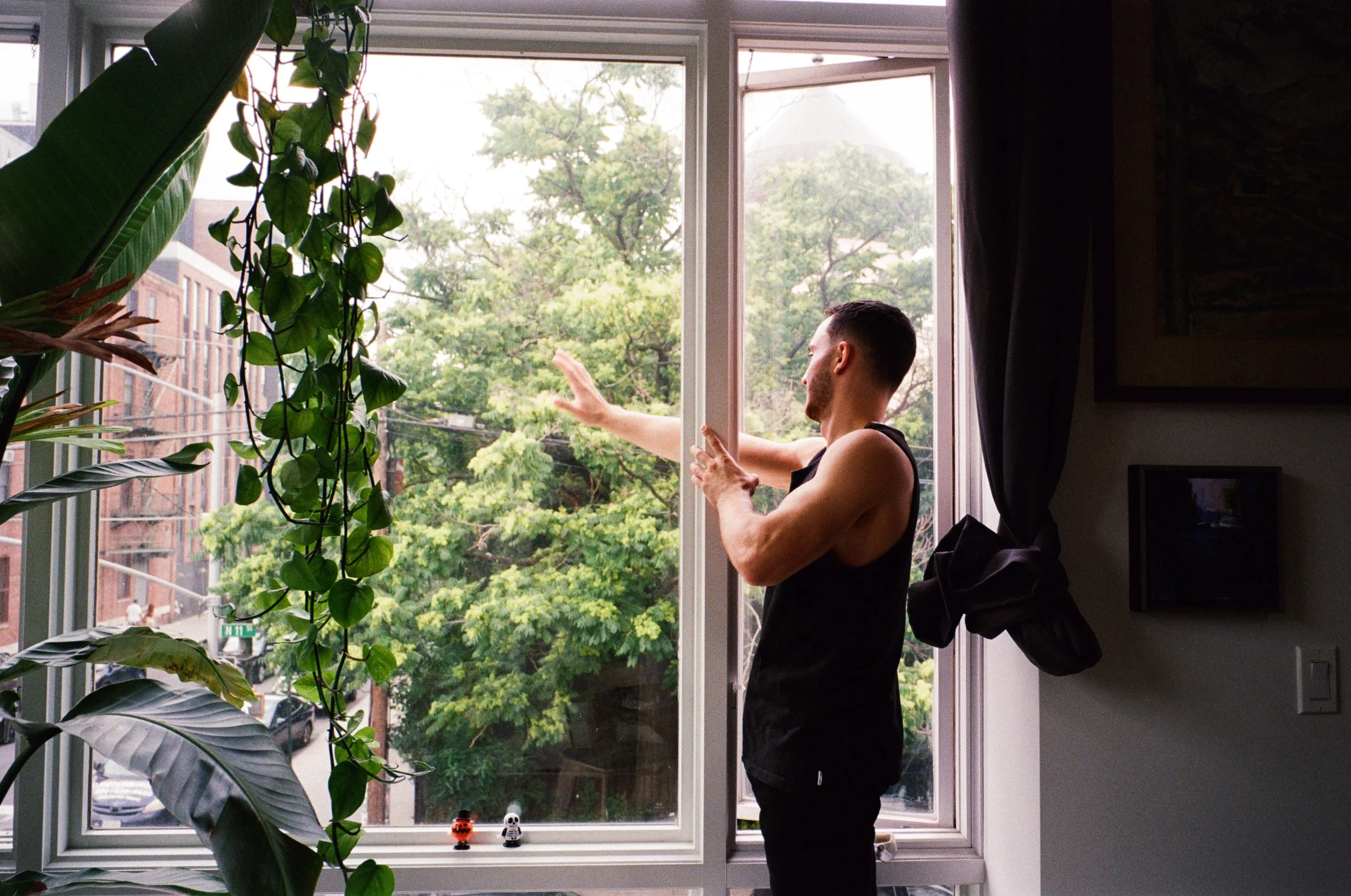 A man in a sleeveless shirt standing inside, reaching out to open a window, with green trees outside and plants inside the room.