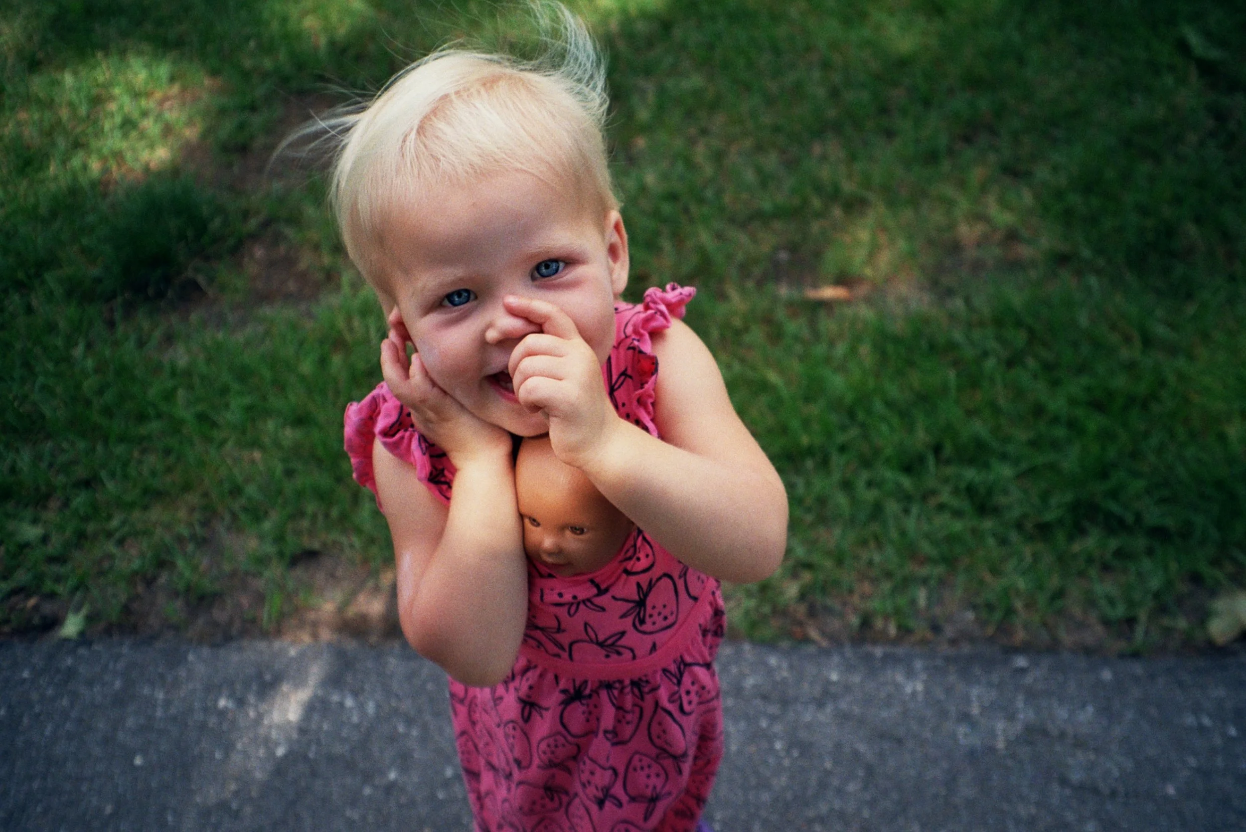 A young girl with blonde hair and blue eyes standing outdoors on grass and pavement, smiling and holding a small doll in her pink dress.