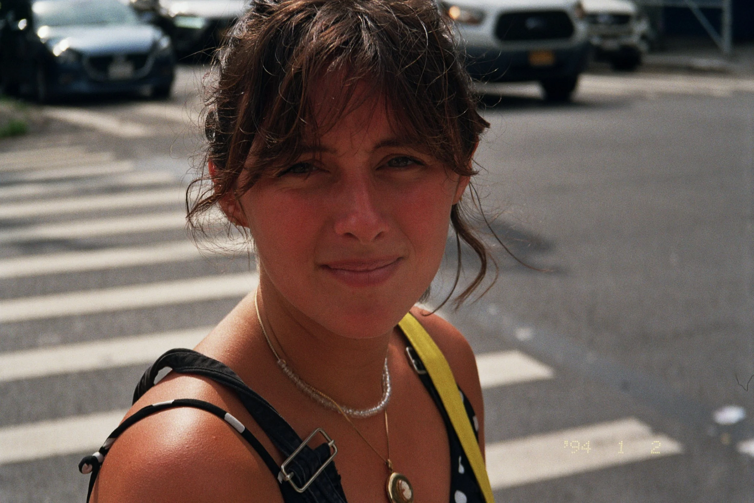 A woman with short, wavy brown hair is looking at the camera while standing outdoors near a crosswalk. She is wearing a black and white polka dot top, a yellow strap, and jewelry including a necklace and a pendant. She has a slight smile and appears 
