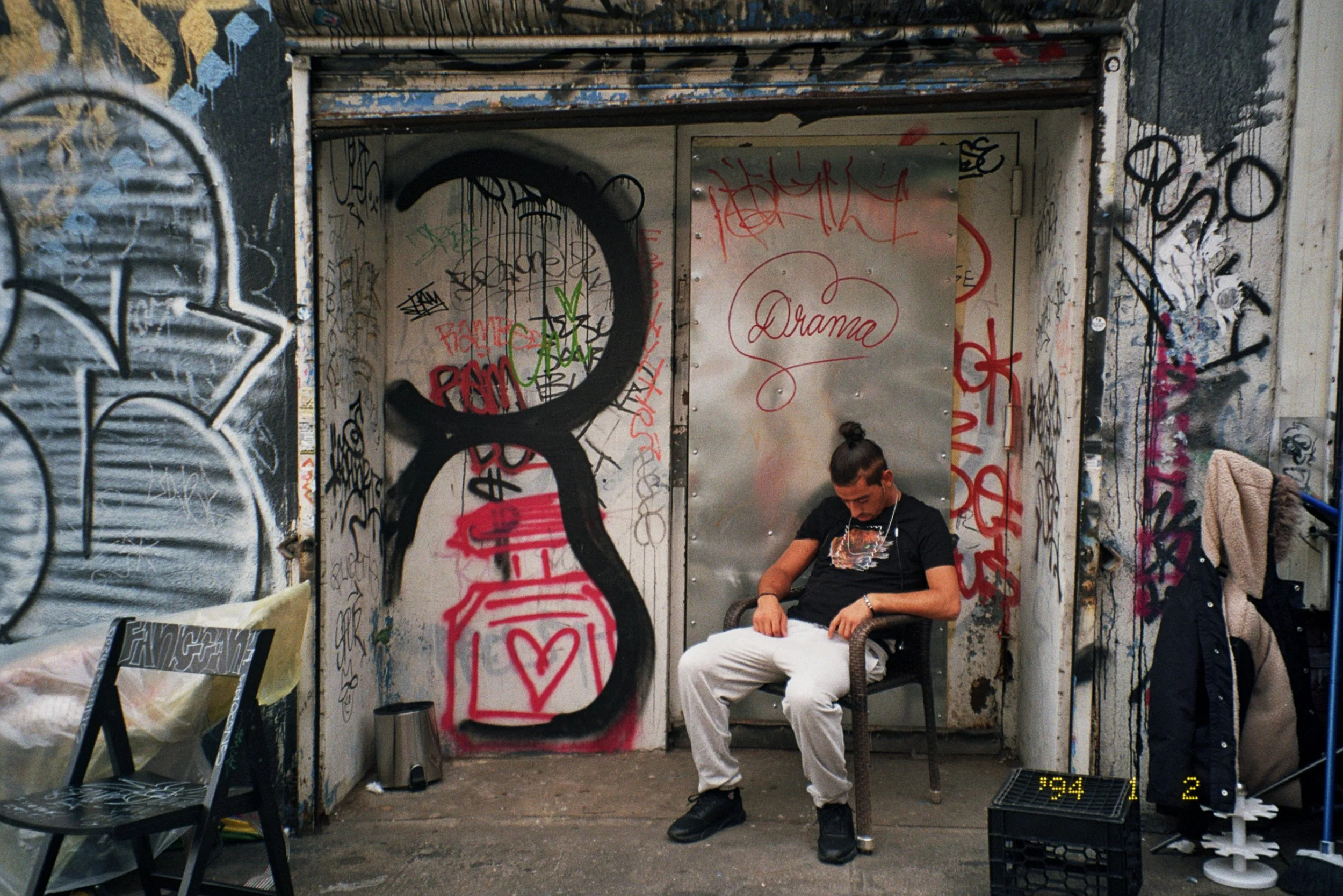 A young man sitting in a graffiti-covered alleyway or bus stop with graffiti on the walls and door, seated on a chair, wearing a black shirt and white pants, looking down at his phone.