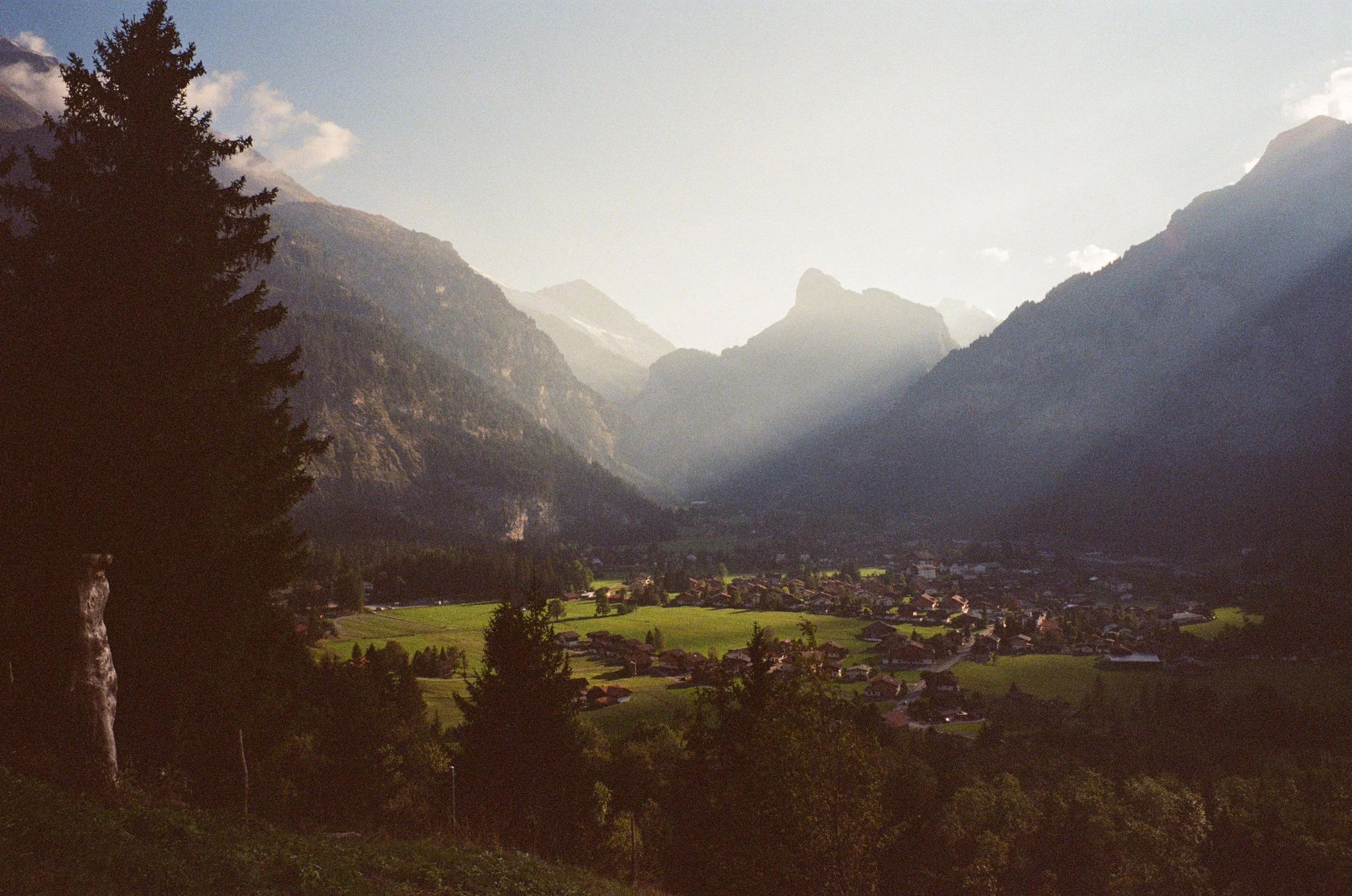 A small village in a green valley surrounded by tall mountains with sunlight streaming through the clouds.