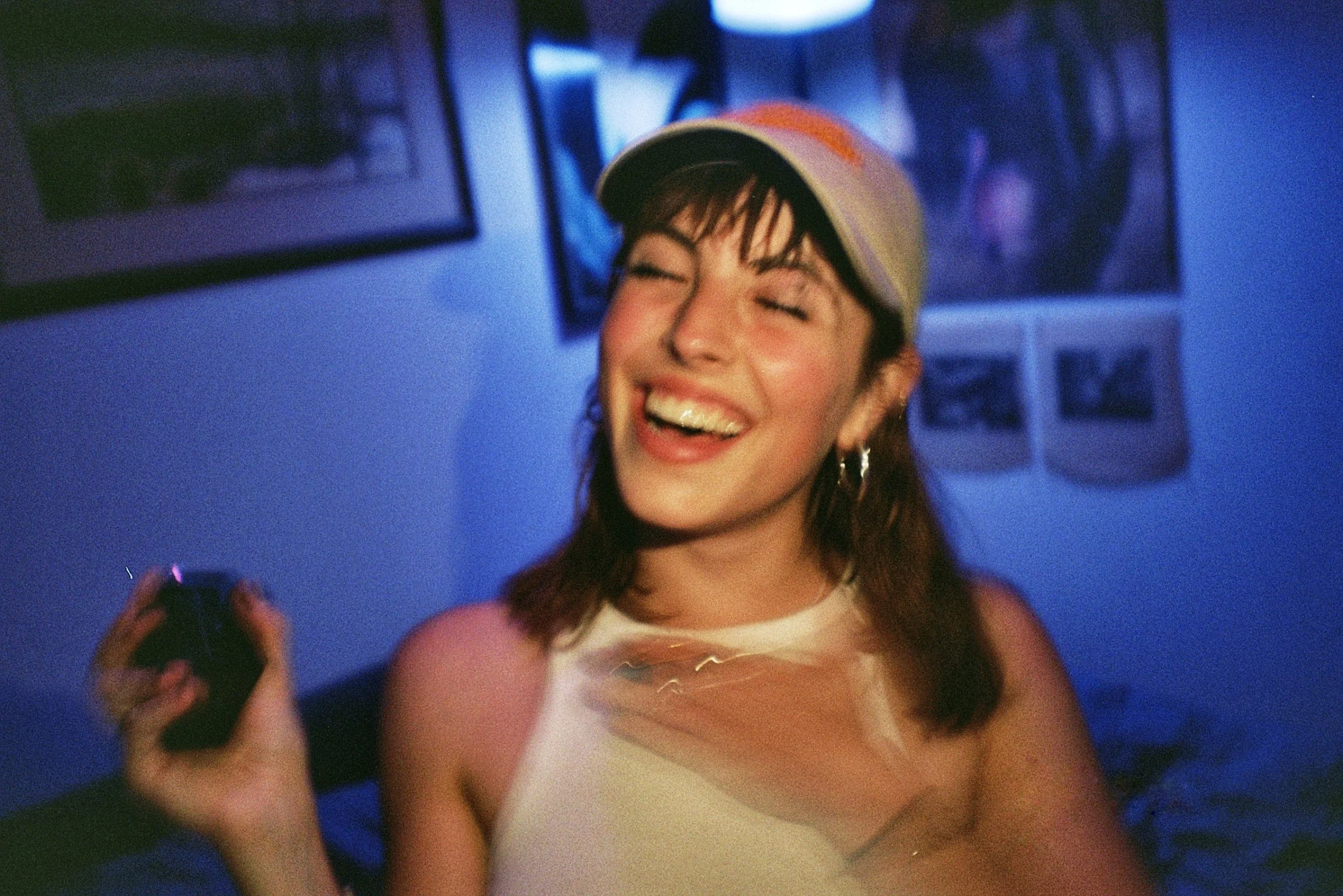 A young woman with brown hair, wearing a yellow and orange baseball cap and a light-colored tank top, laughing joyfully in a dimly lit room with blue lighting and framed pictures on the wall.