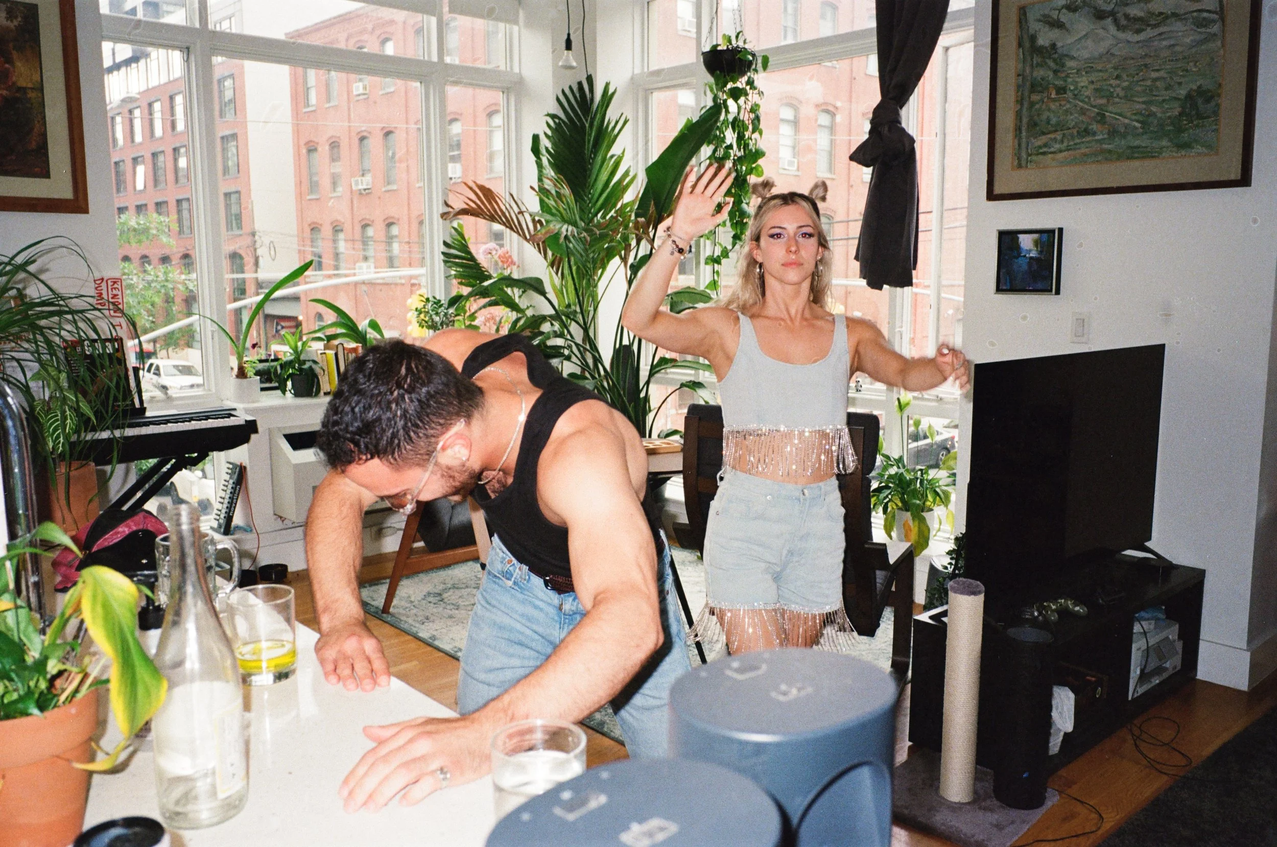 A woman standing with her right arm raised and her left hand extended, inside a living room with large windows, plants, and artwork. A man with glasses and dark hair, wearing a black tank top, is leaning over a white table, seemingly drinking or reac