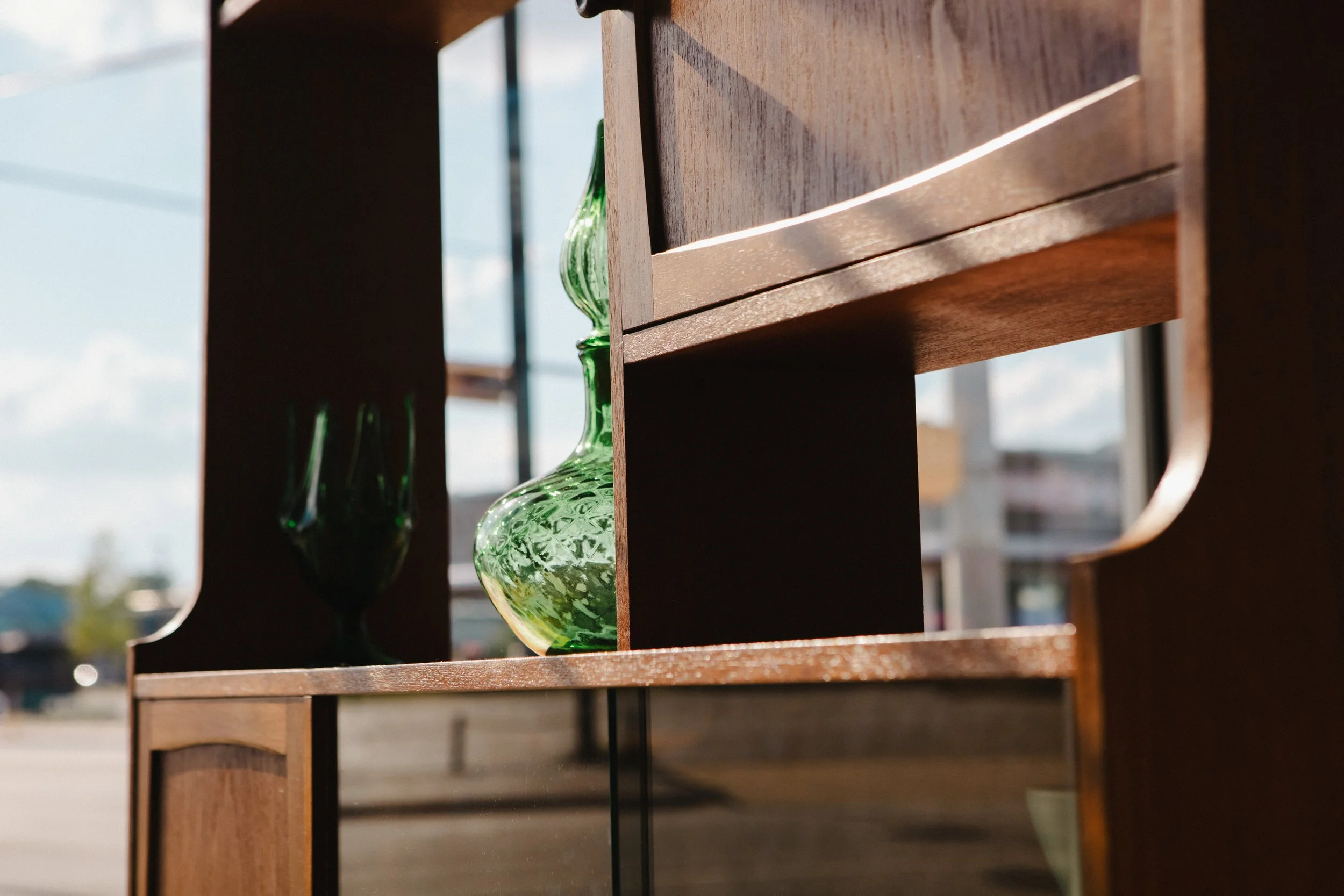 Close-up of a wooden shelf with a green glass vase and a dark glass vessel, with a window and blue sky in the background.