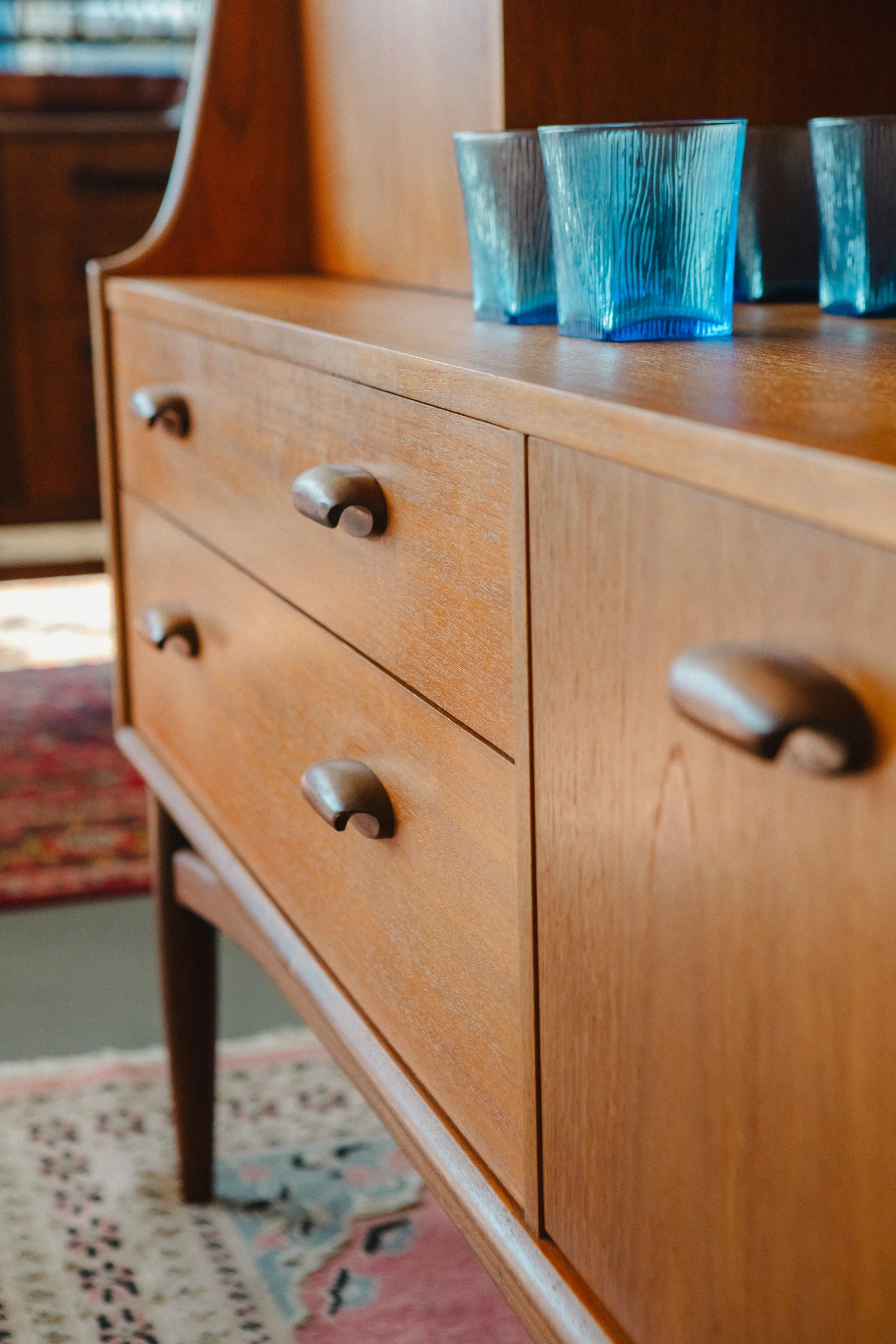 A wooden dresser with four curved knobs, on top of which are four blue glass cups, in a room with a patterned rug and other wooden furniture.