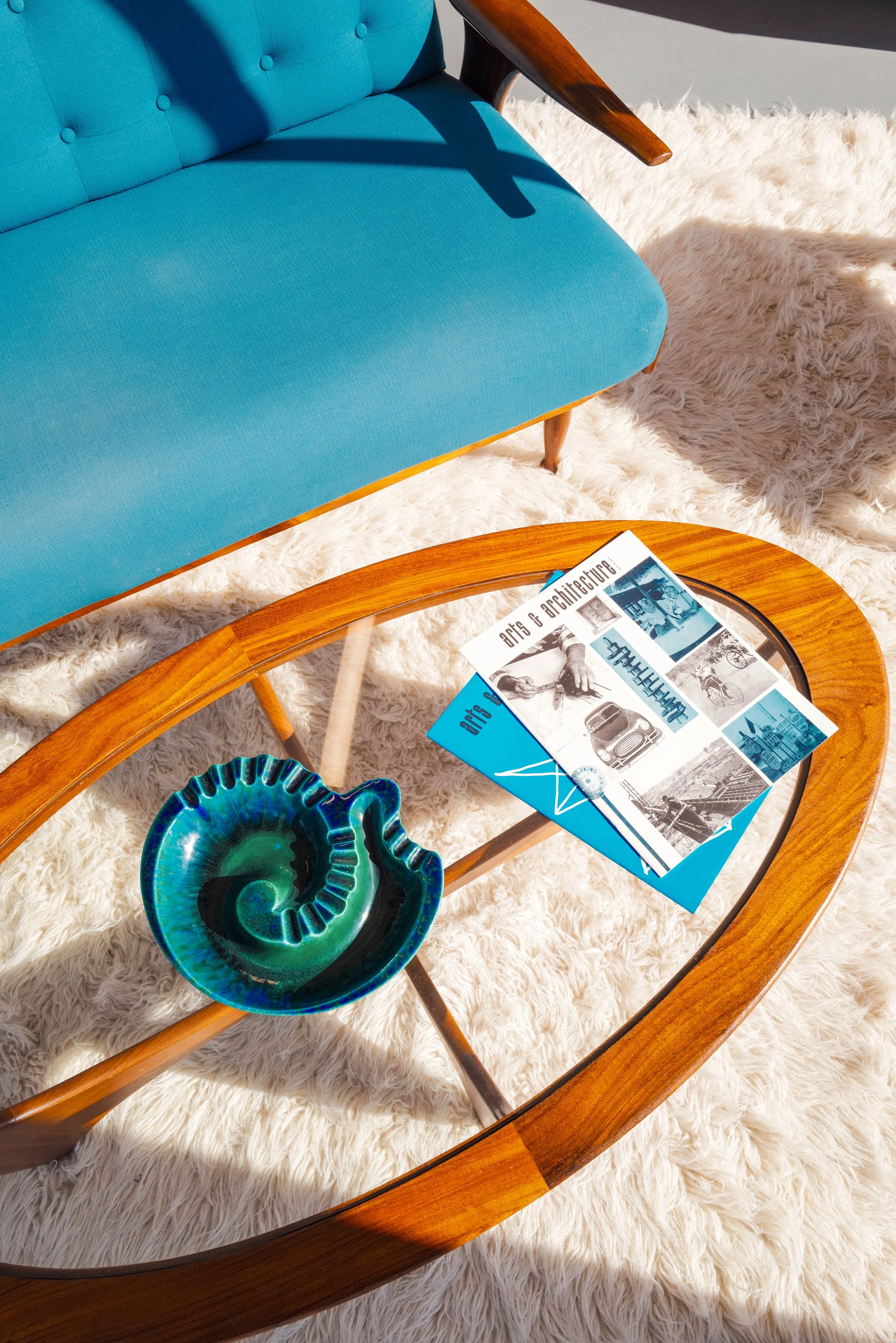 A wooden coffee table with a glossy turquoise ceramic ashtray and a brochure or magazine on top, next to a mid-century style blue sofa and a white shag rug.