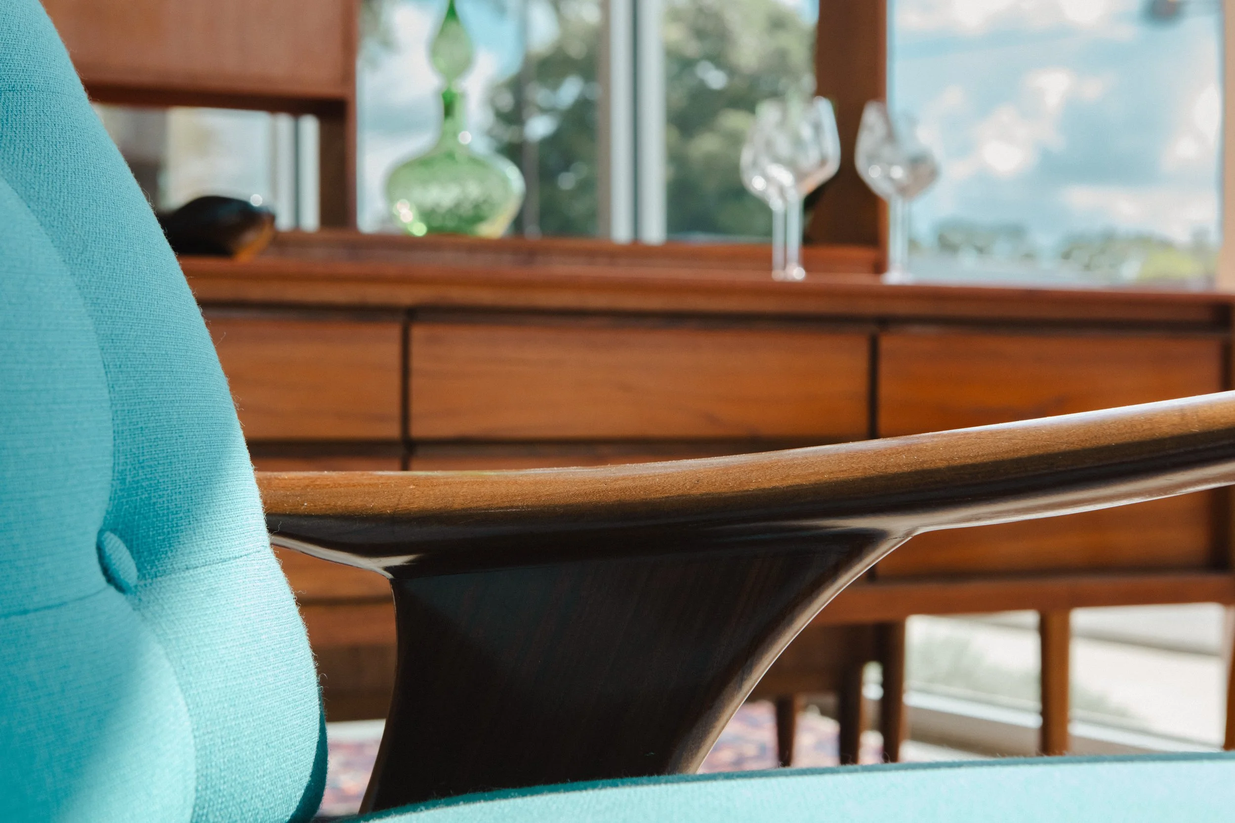 Close-up of a turquoise upholstered chair with a wooden armrest, with a wooden dresser and decorative glass items in the background.