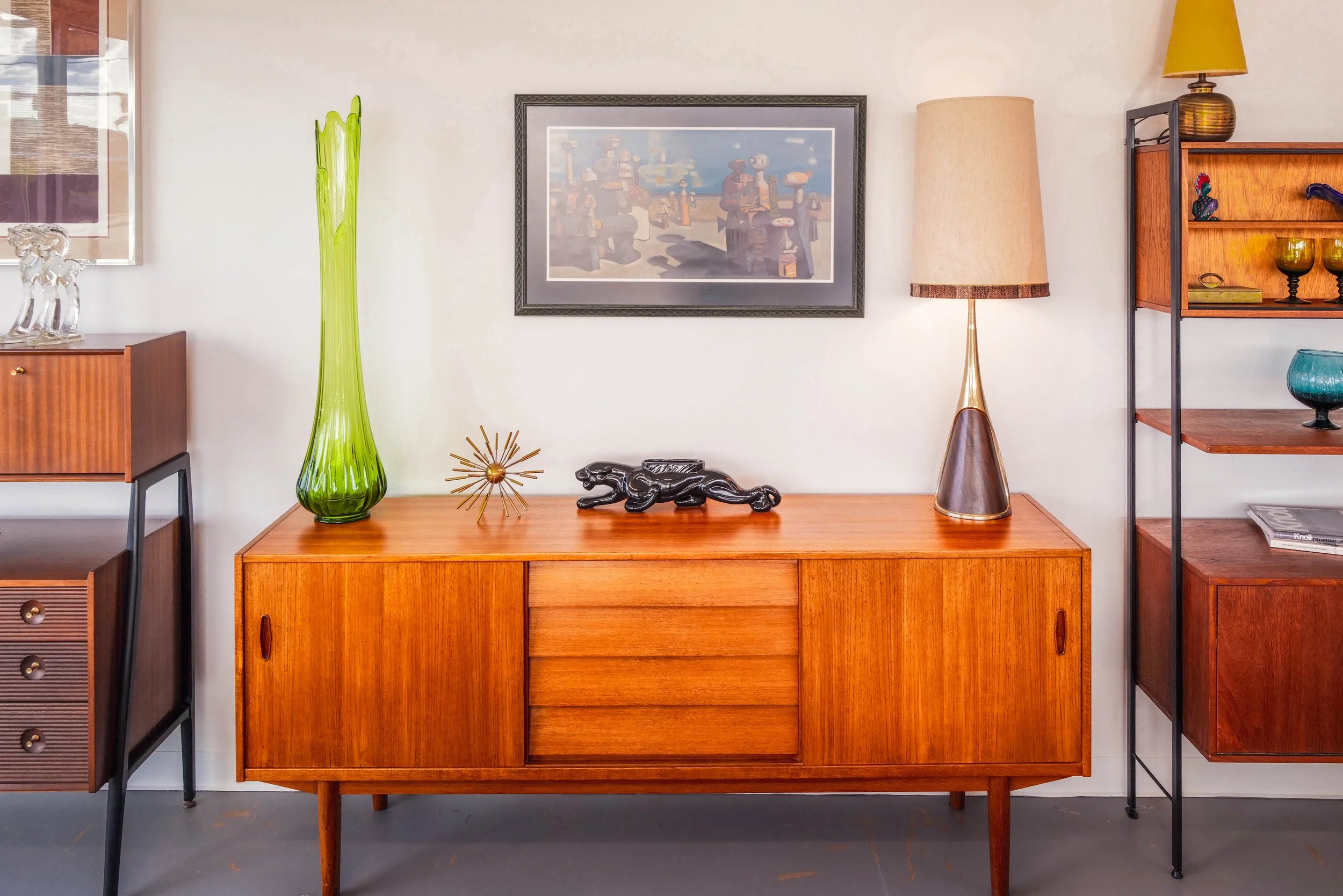 Mid-century modern wooden sideboard with decorative items, including a tall green glass vase, black ceramic horse sculpture, a gold starburst ornament, and two lamps, one beige and one yellow, on a wall with framed artwork and a white wall.
