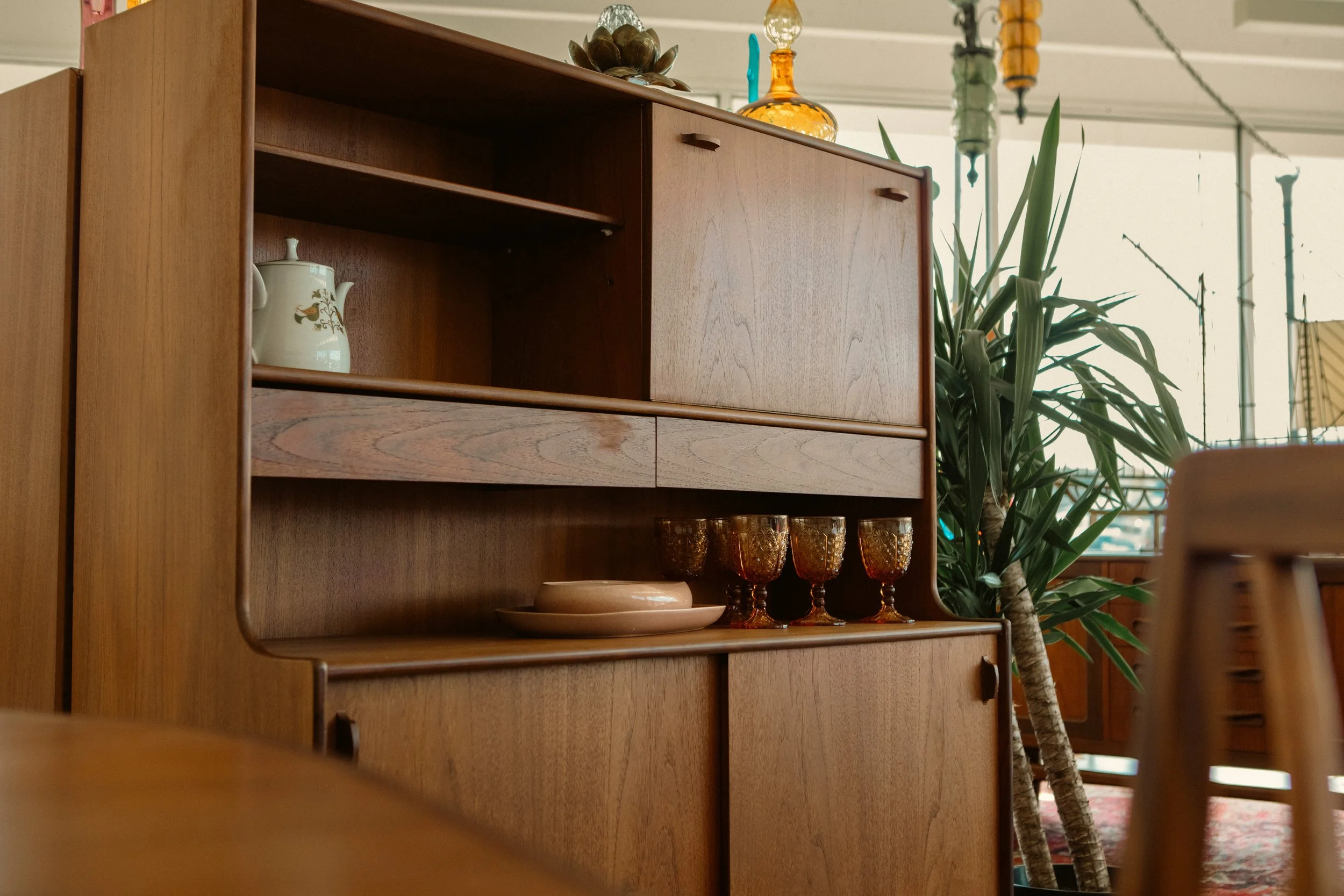 A wooden cabinet with decorative glassware and a teapot, situated next to a potted plant in a well-lit indoor space.