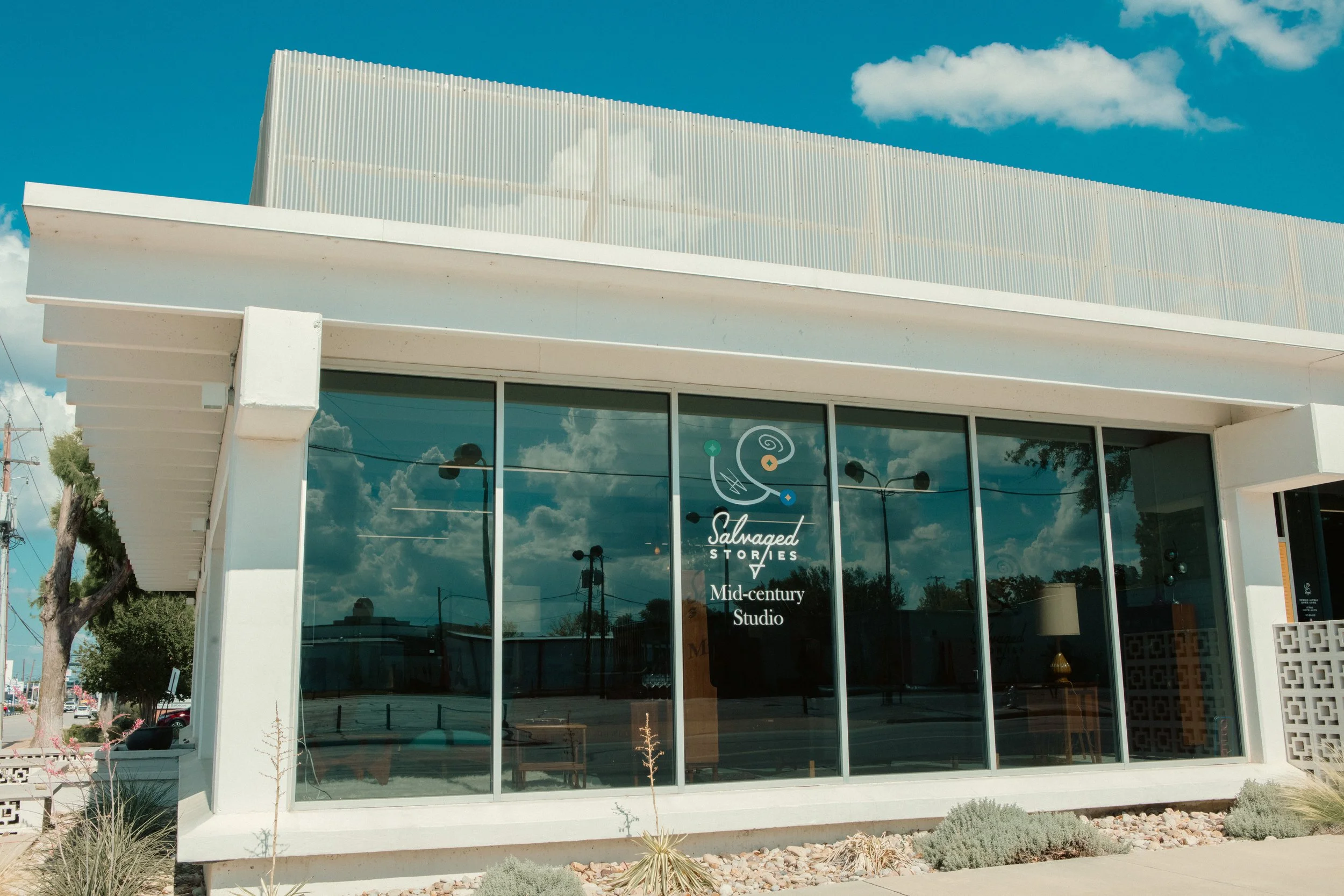 Front view of a modern building with large glass windows and white exterior, reflecting a blue sky with clouds, trees, and power lines. The building has a logo and text on the window that reads 'Salvaged Stories, Mid-century Studio.'