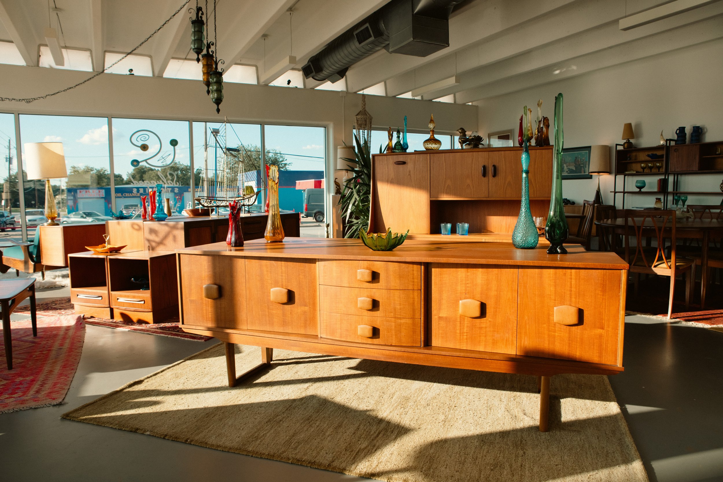 Interior of a vintage furniture store with a large mid-century modern wooden sideboard in the foreground, decorated with colorful glass vases and a small green bowl, and other wooden furniture and decor items in the background, with large windows allowing natural light inside.