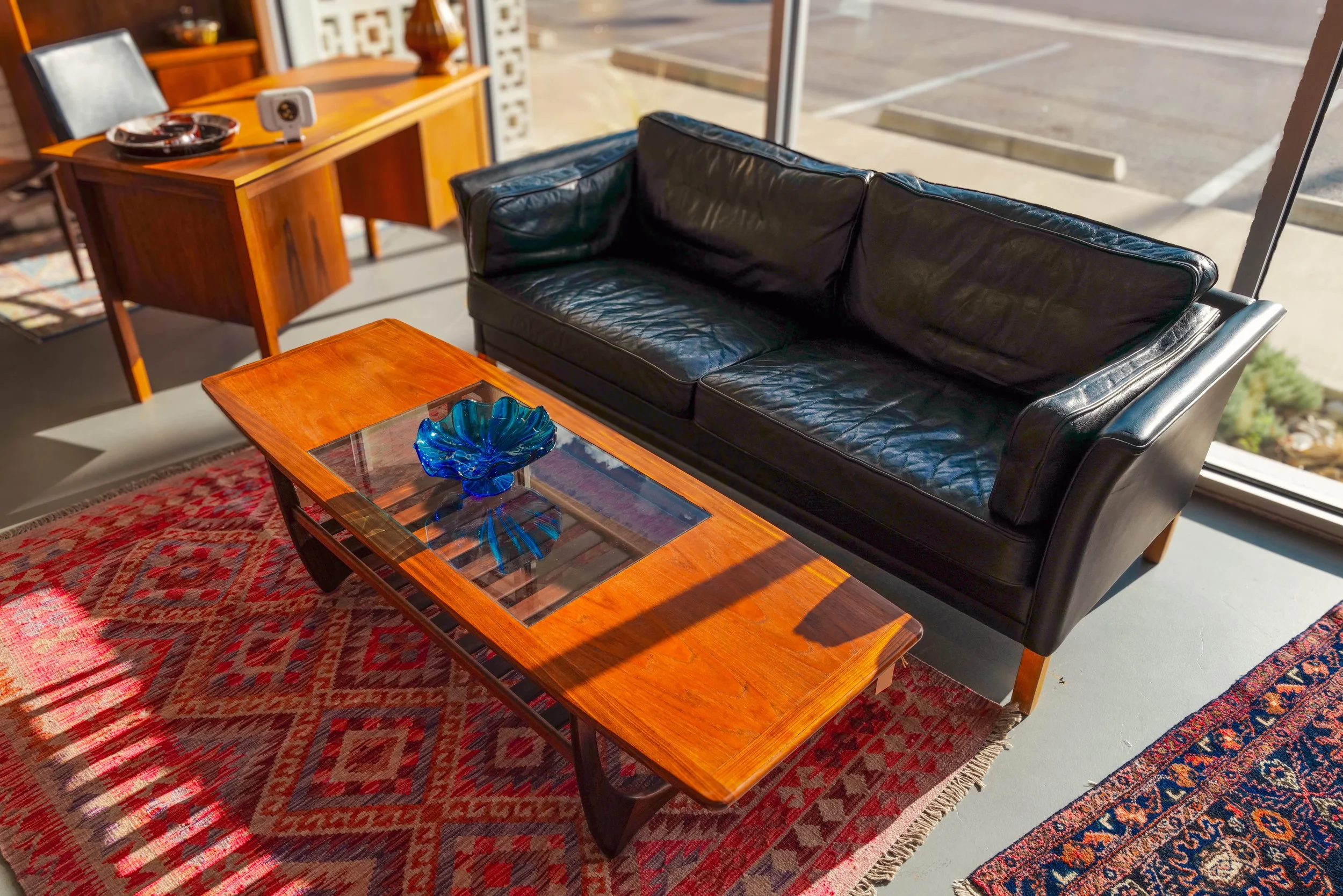 A living room with a black leather sofa, a wooden coffee table with a blue glass bowl on top, a red patterned rug, and a wooden side table in front of a large window showing a parking lot outside.