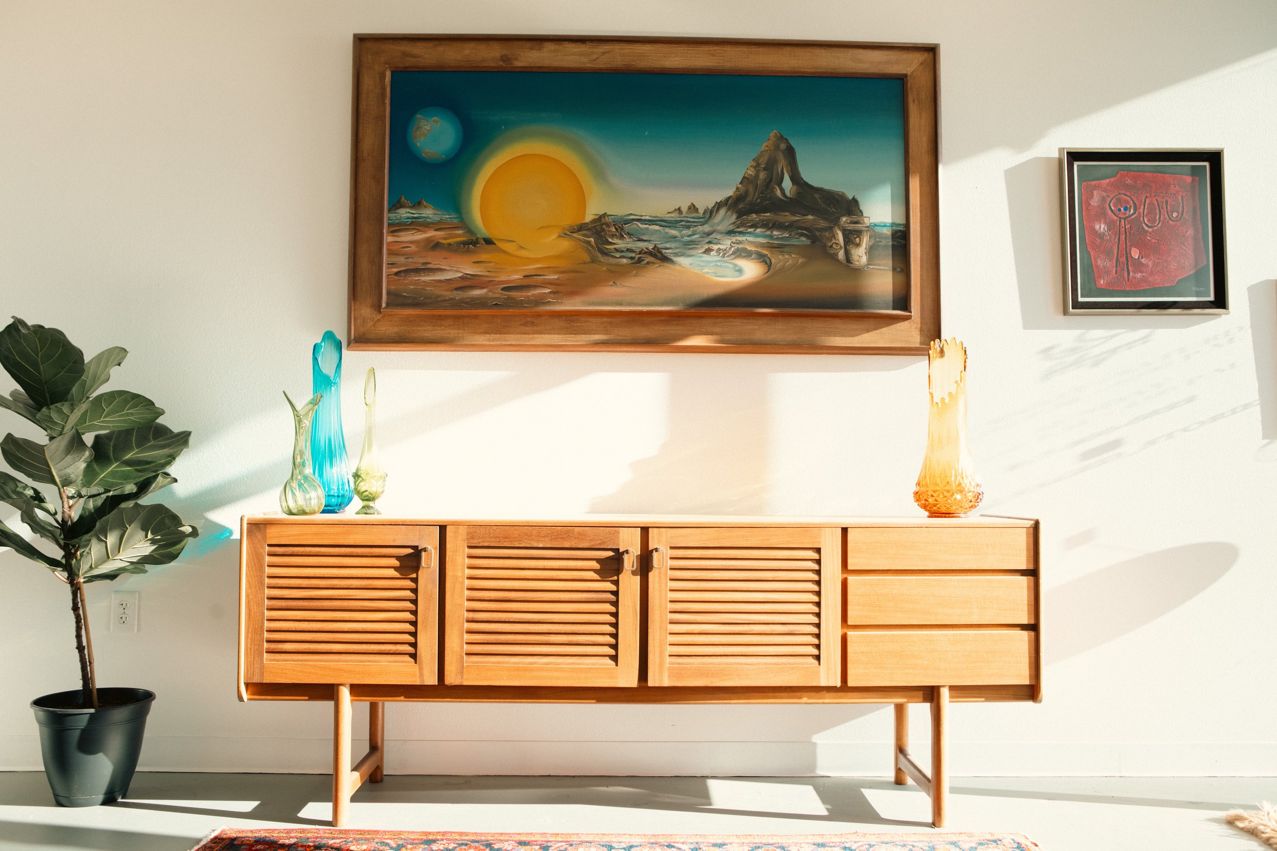A mid-century modern wooden sideboard decorated with glass vases placed on top. Behind it is a white wall adorned with a large landscape painting of a desert scene with a sun, mountains, and a rock formation, in a wooden frame. To the right is a small framed abstract artwork. To the left, a large potted green plant. Sunlight creates shadows on the wall.