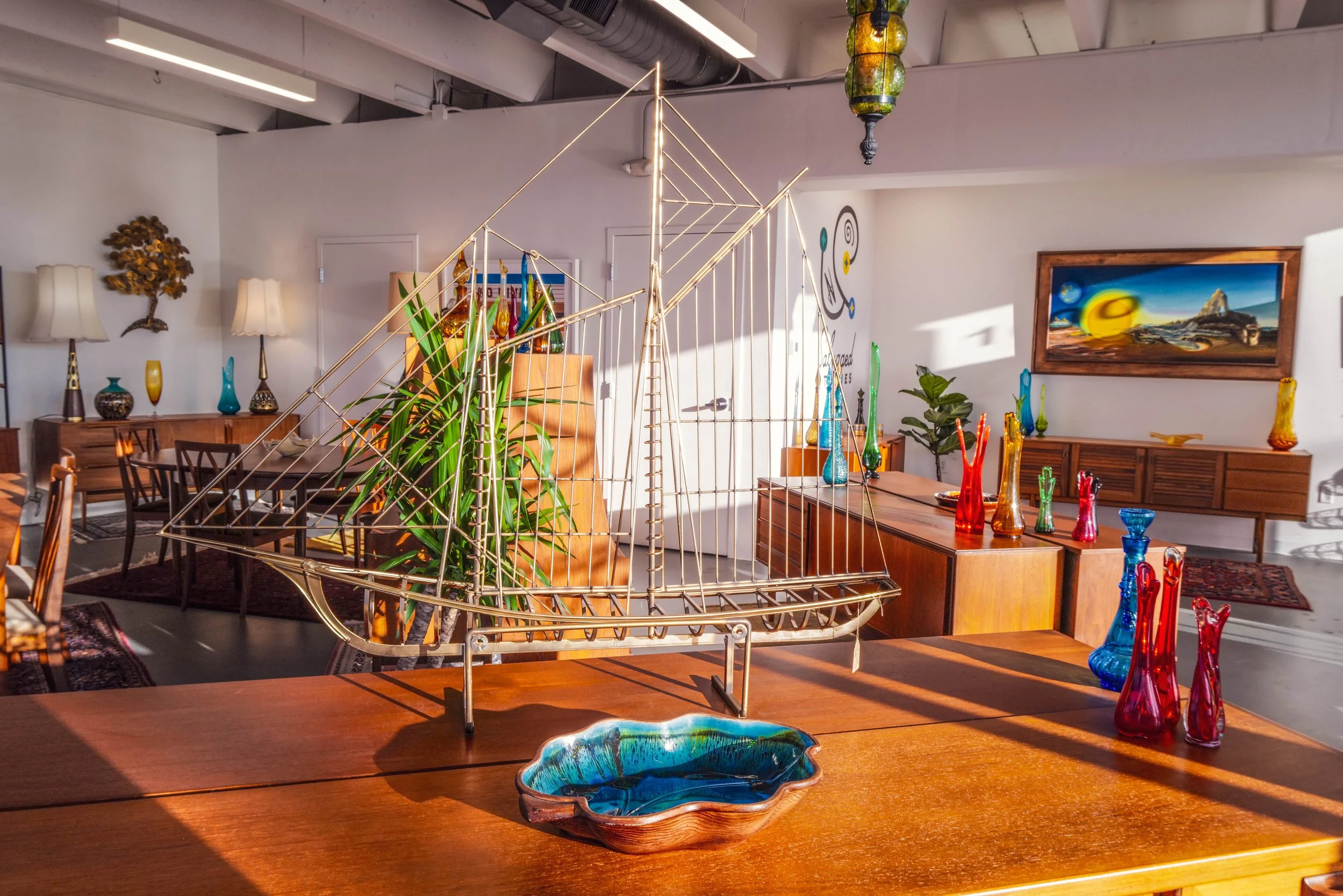 A souvenir metal sailboat model and a decorative ceramic bowl on a wooden table in a room decorated with colorful vases, paintings, and plants.