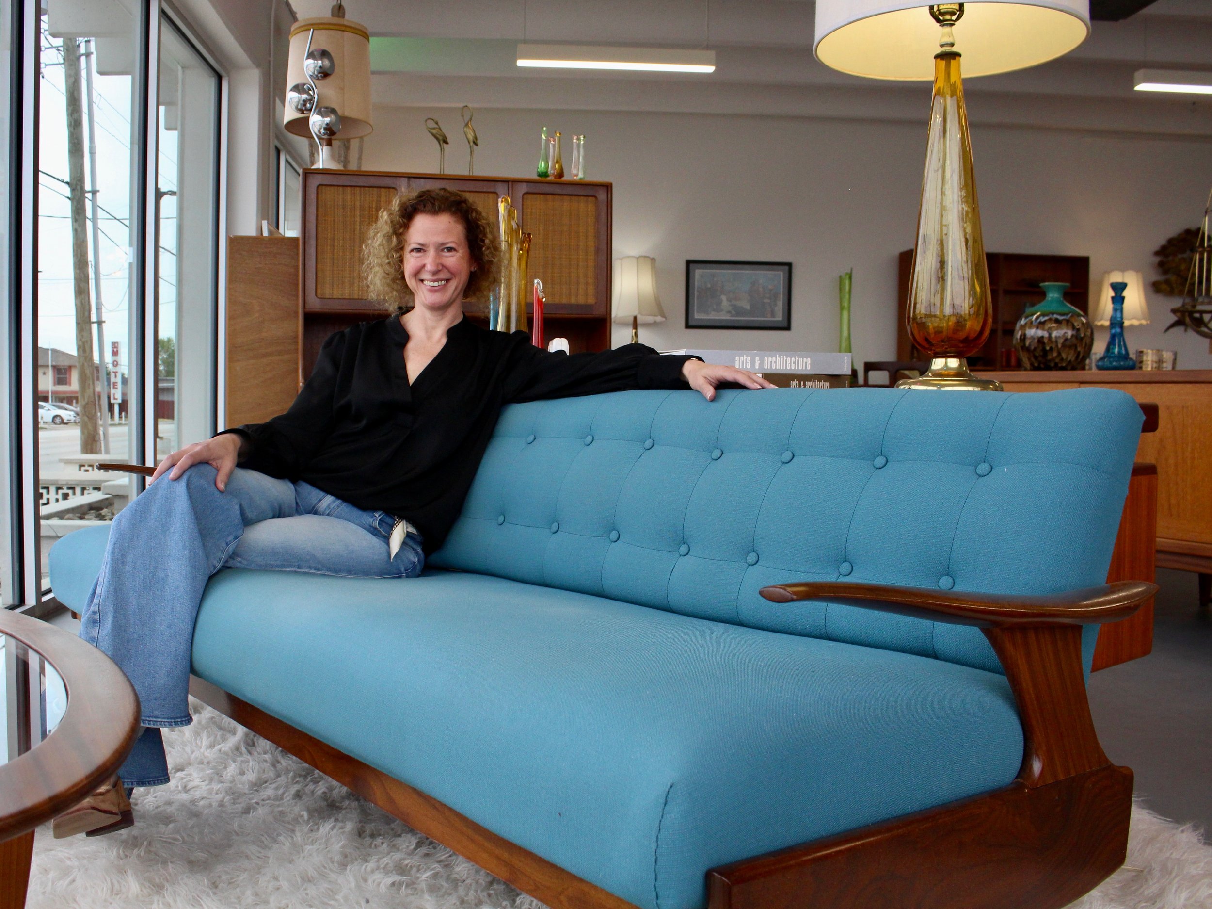 A woman with curly hair wearing a black top and jeans sitting on a mid-century modern blue couch inside a furniture store.