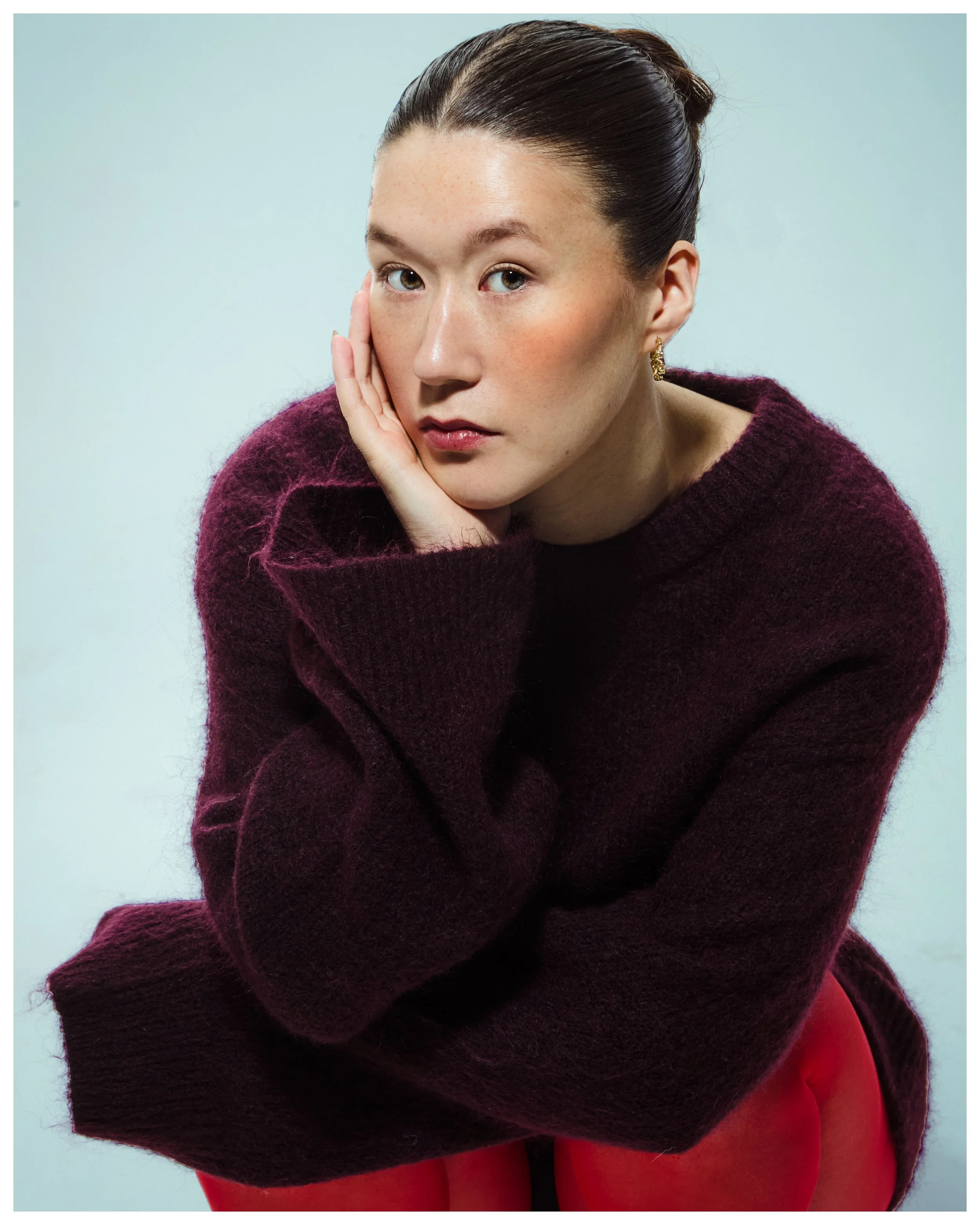 A woman with dark hair in a bun, wearing a maroon sweater and red pants, resting her head on her hand, looking at the camera against a light background.