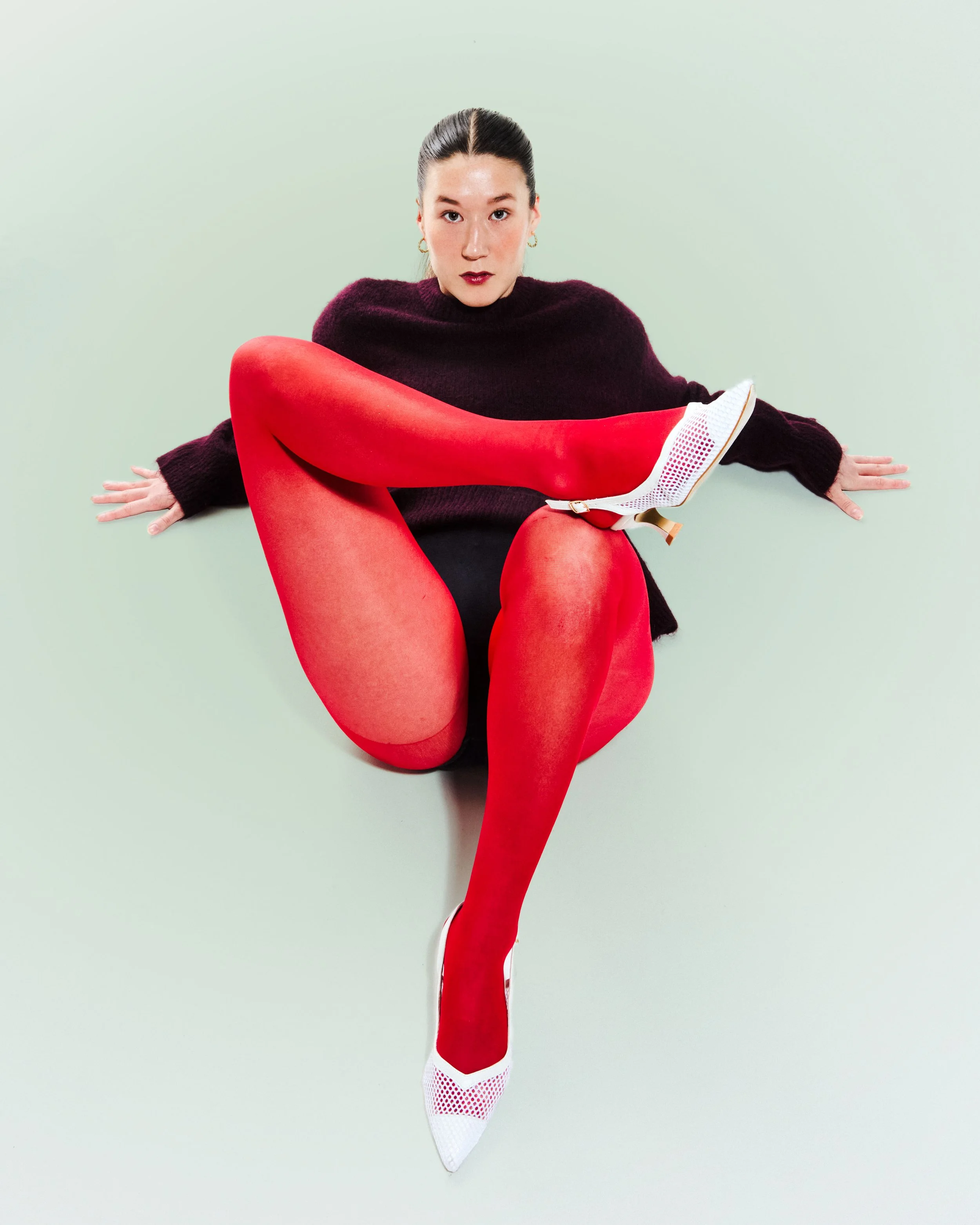 Woman wearing red tights, black shorts, dark sweater, and white heels sitting on the floor against a pale green background with her legs crossed.