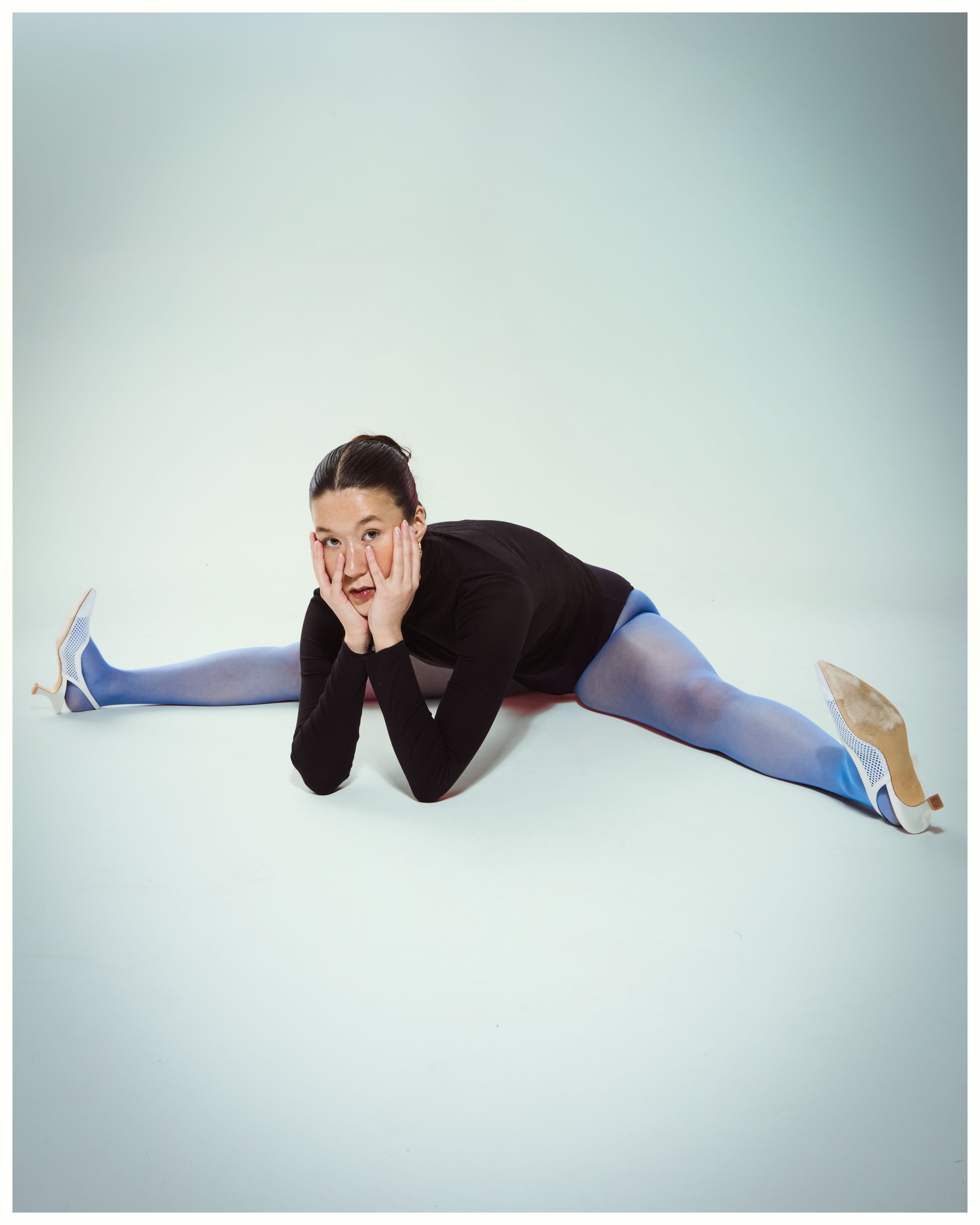 A woman lying on the floor in a side split pose, resting her head on her hands, wearing a black top, blue tights, and high heels, against a plain white background.