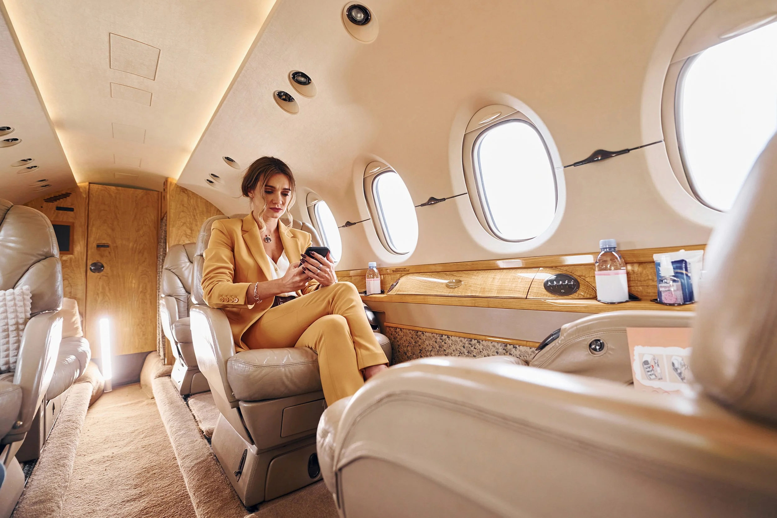 A woman in a yellow suit sitting on a private jet, using her phone, with windows and beige leather seats visible.