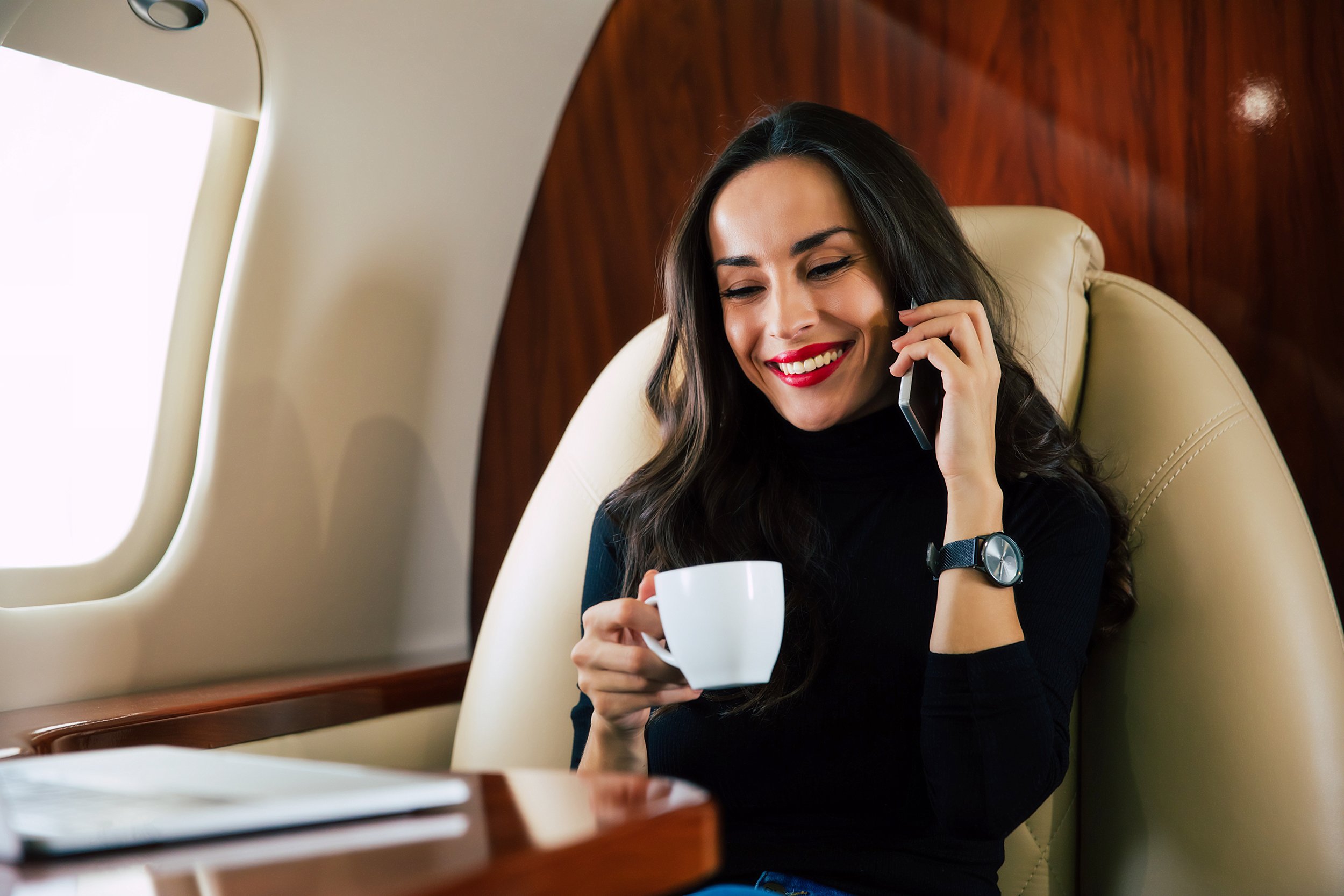 A woman with long dark hair, wearing a black top, smiling, holding a white mug in one hand, and talking on a phone with the other hand, seated inside a private jet.