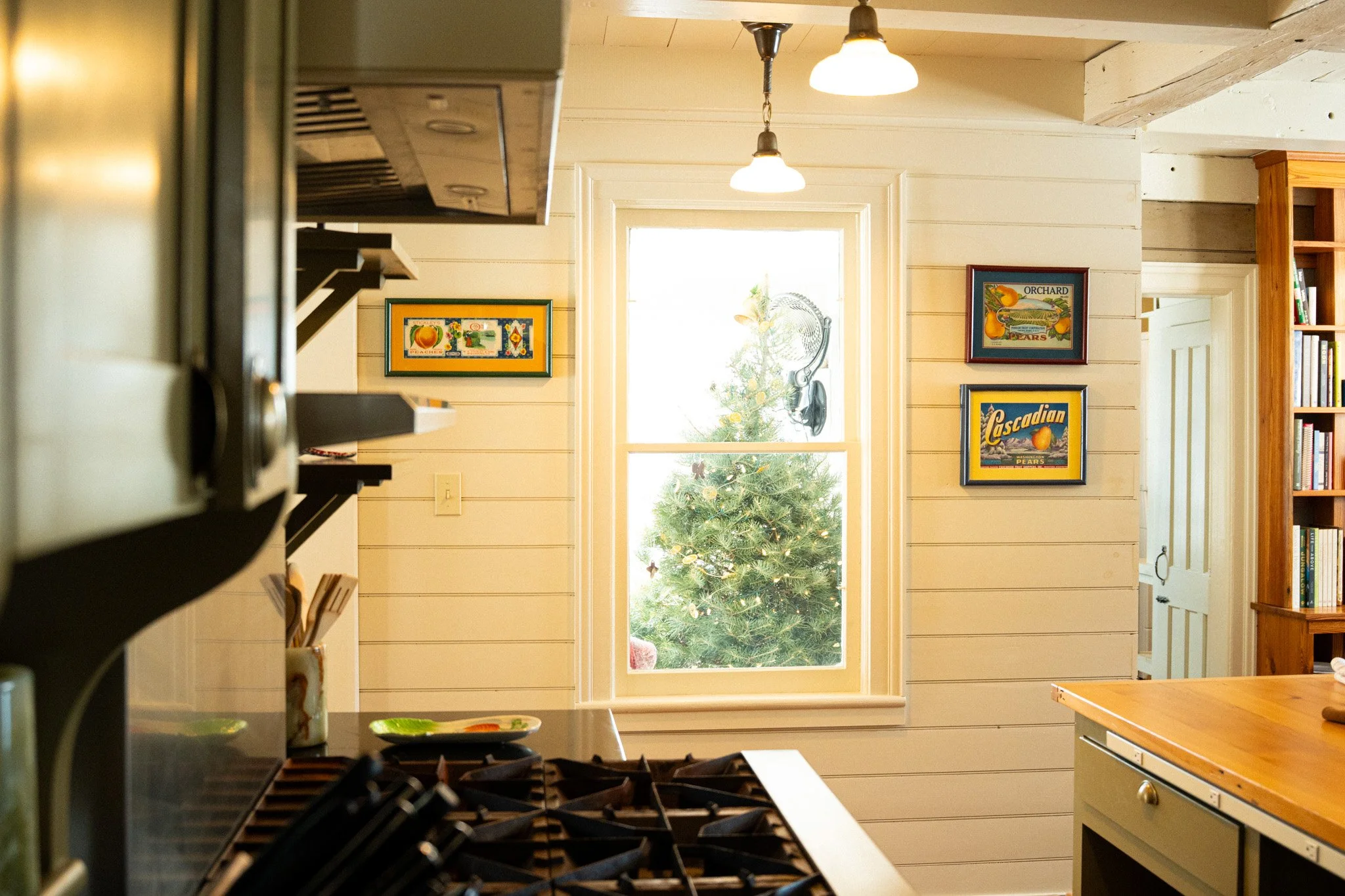 Kitchen with a window showing a decorated Christmas tree outside. Decorated wall with colorful paintings, wooden shelves, and a wooden countertop.