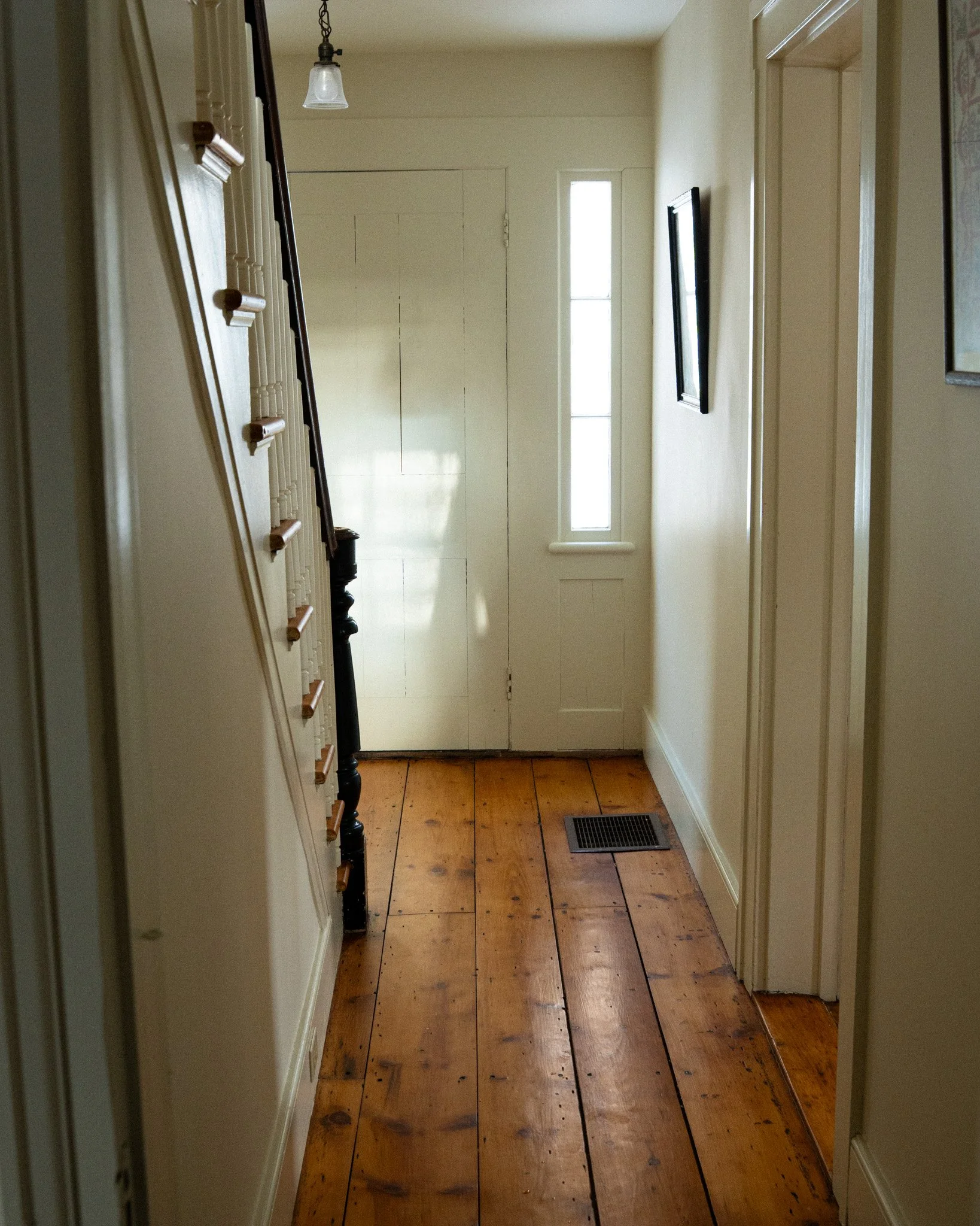 Foyer with wooden floor, white walls, a narrow vertical window, a front door, hanging light fixture, and framed artwork.