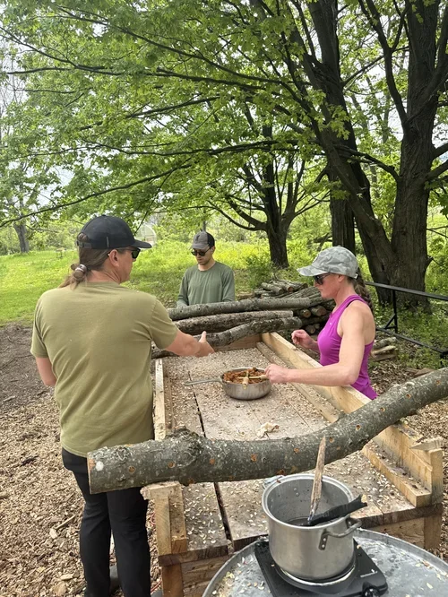 Three people outdoors constructing a wooden structure with logs, surrounded by green trees and grass.