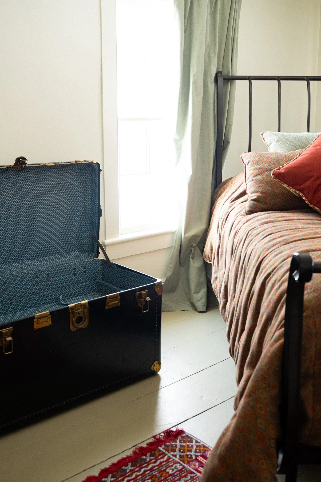 An open vintage black suitcase on the floor next to a bed with a patterned quilt and assorted pillows, located near a window with light green curtains in a bright room.