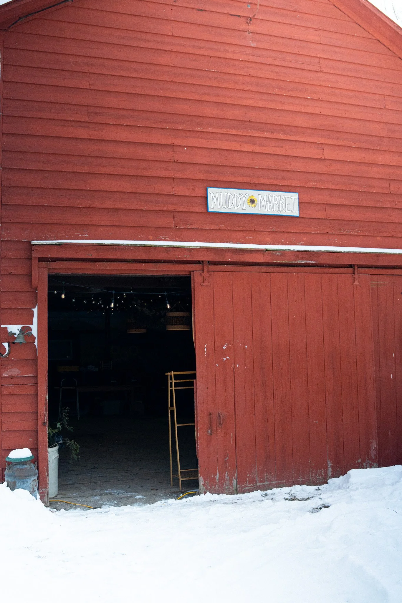 Red barn with open sliding door, snow on ground, and sign that reads 'Muddy Market' with a sunflower illustration.
