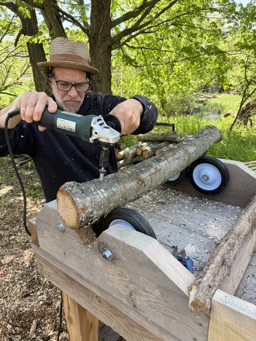 A man wearing a straw hat, glasses, and a black shirt using a power tool on a large tree branch outdoors in a green, wooded area.