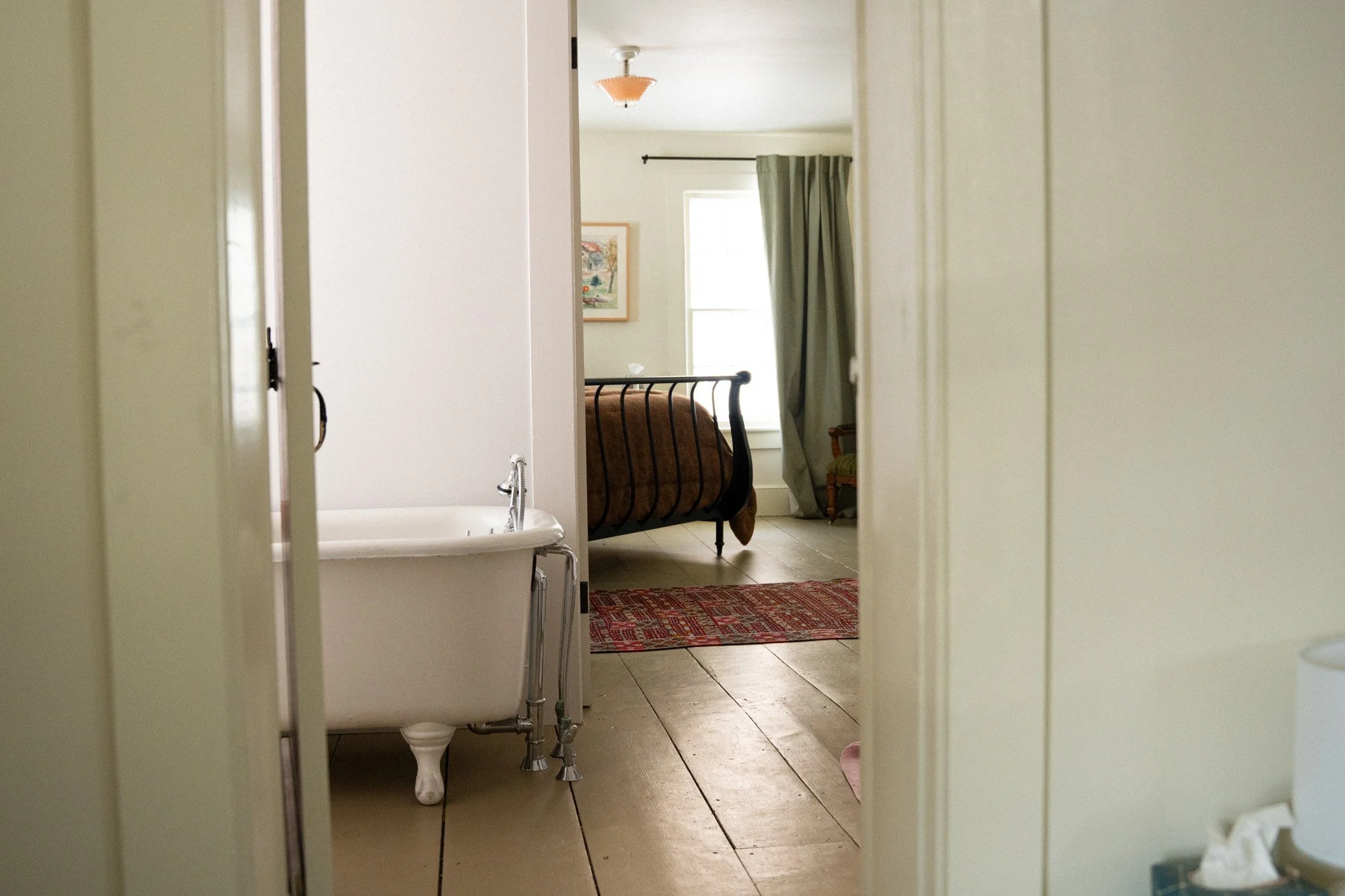 View of a bedroom through an open doorway, featuring a vintage clawfoot bathtub in the foreground, a bed with a metal frame, a window with green curtains, and a red patterned rug on the wooden floor.