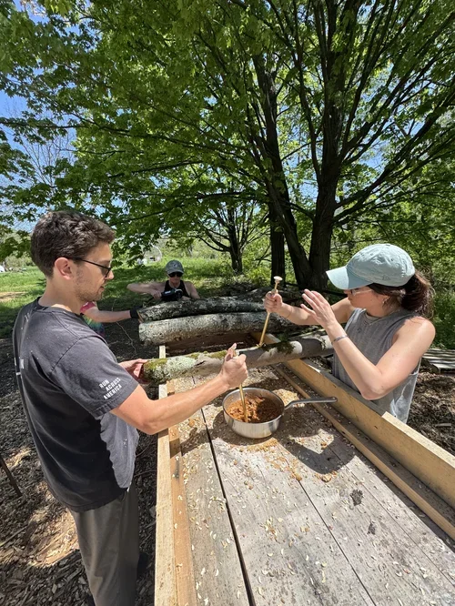 Two people are working on a woodworking project outdoors, surrounded by trees, with a small child in the background watching.