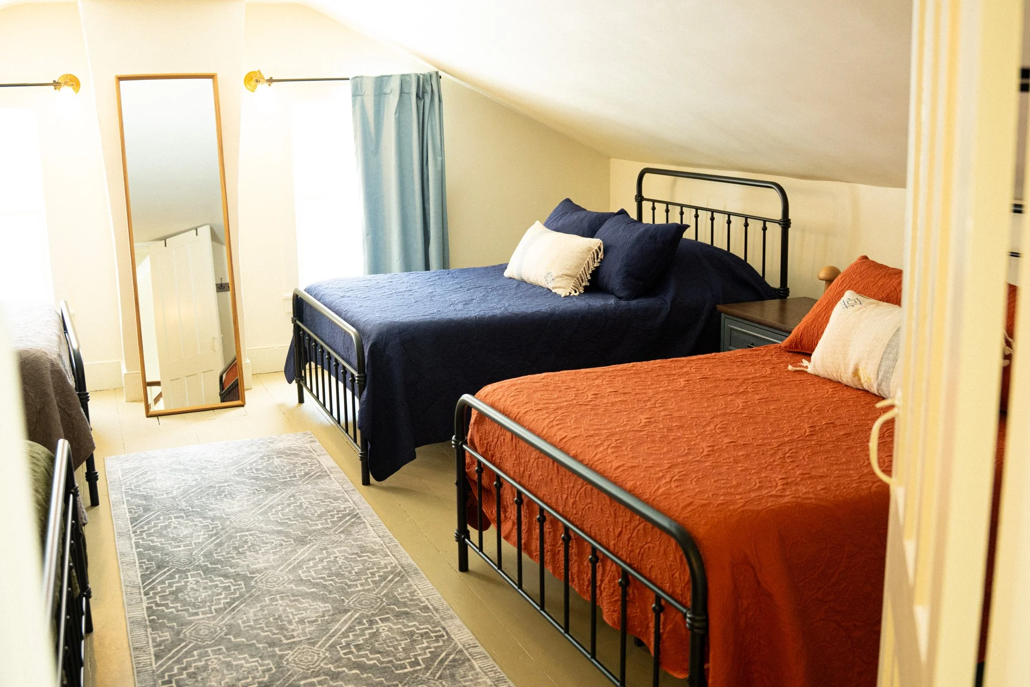A cozy bedroom with two metal beds; one with a navy blue quilt and pillows, the other with an orange quilt and pillows. A full-length mirror leans against the wall next to a window with light blue curtains, and a gray patterned rug covers part of the