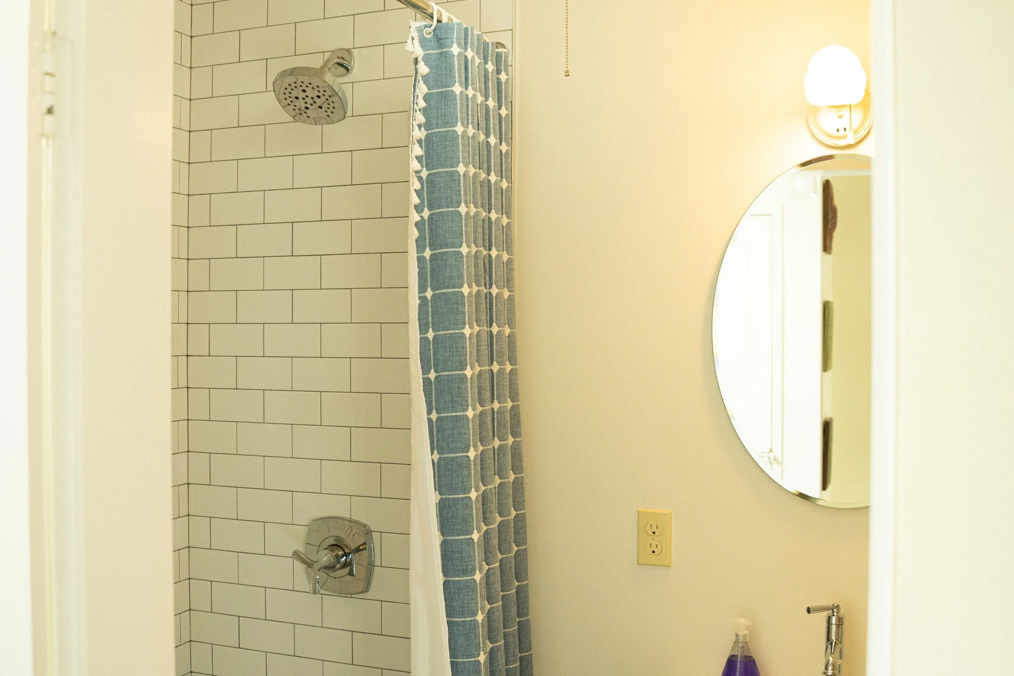 A bathroom with a shower area featuring white subway tiles, a silver showerhead, and a blue patterned shower curtain. To the right is a wall with a round mirror, a wall-mounted light fixture, and a partially visible sink with a purple soap dispenser.