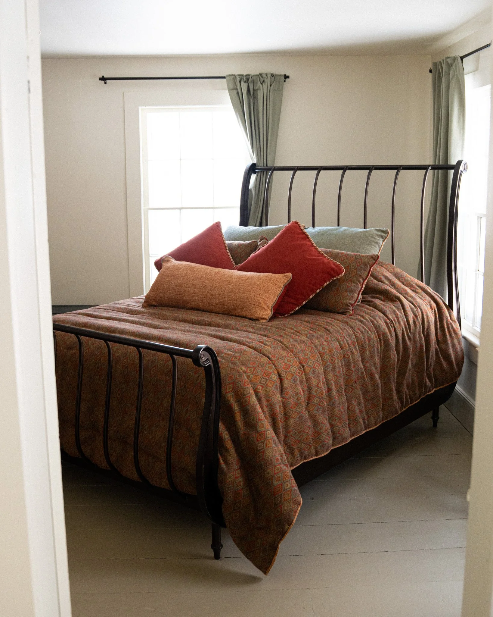 A bedroom with a metal bed frame, covered with a patterned brownish-orange quilt, and decorated with multiple pillows in shades of red, orange, and beige. There are two windows with green curtains, allowing natural light to brighten the room.