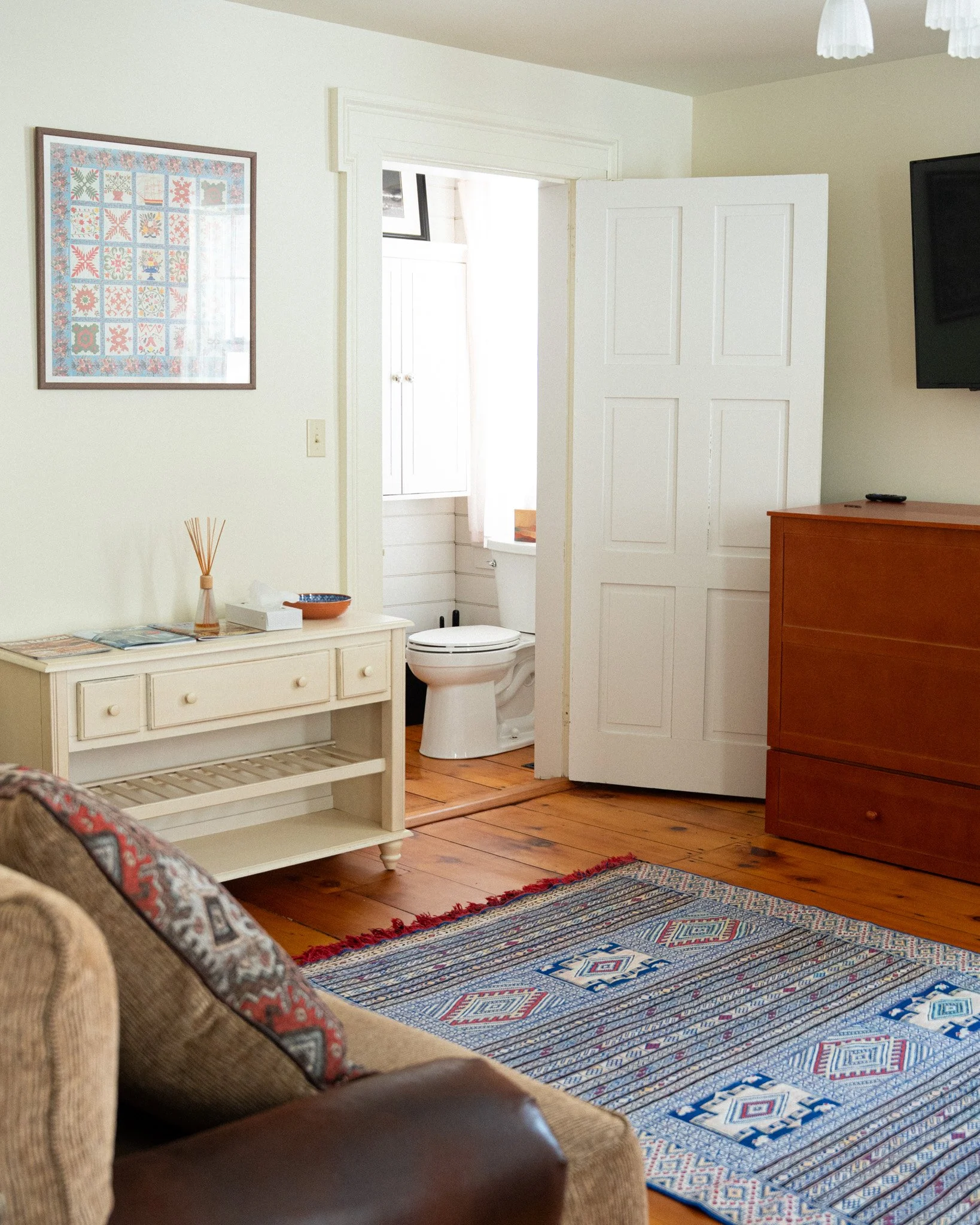View of a cozy living room with a beige sofa, patterned rug, and a white side table next to a doorway leading to a small bathroom with a toilet and window. A wooden dresser and a wall-mounted TV are also visible.