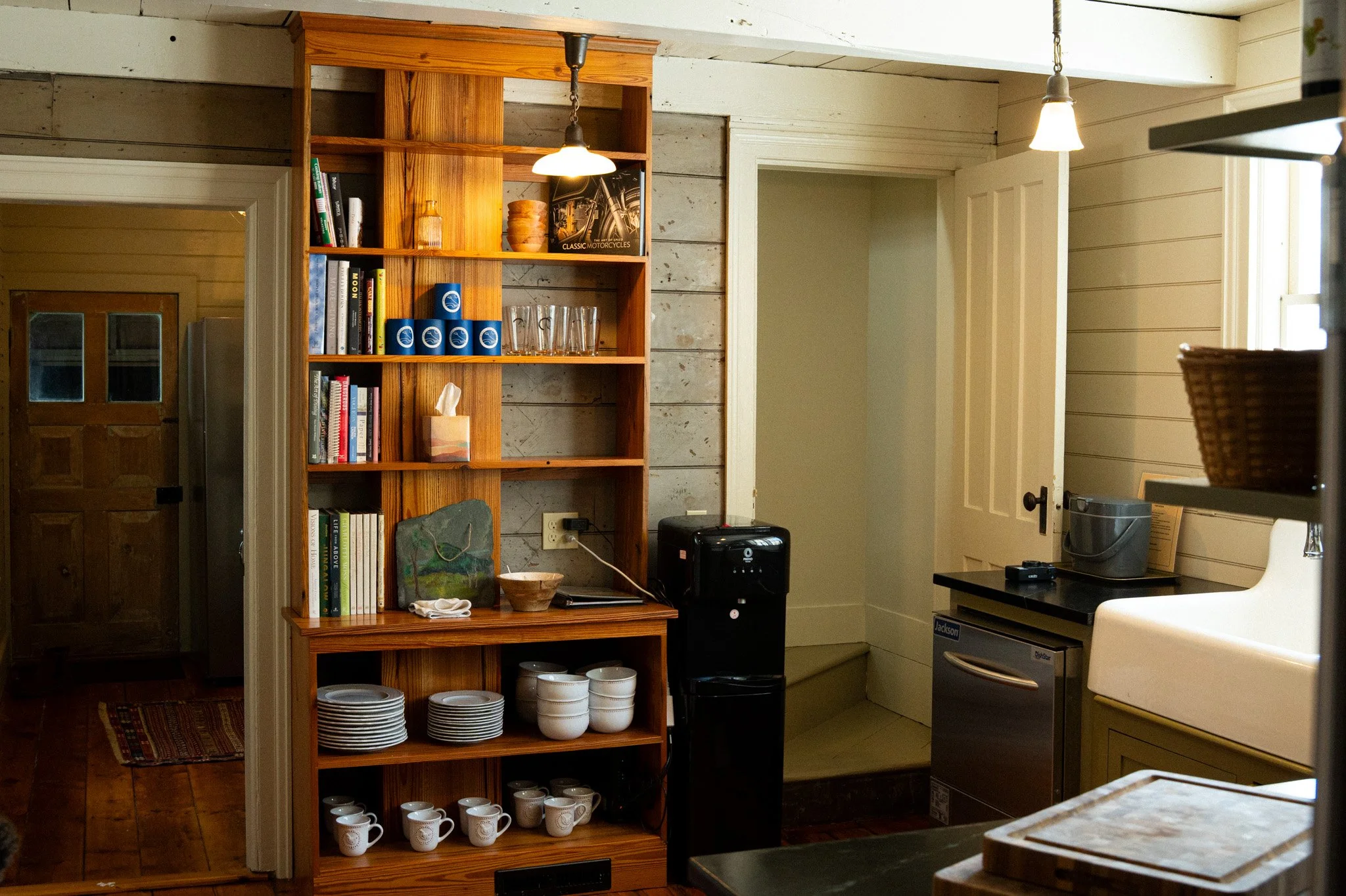 Interior of a cozy kitchen with wooden shelves holding books, glasses, and cups, a sink, a small refrigerator, and a water dispenser, with warm lighting and a rustic decor style.