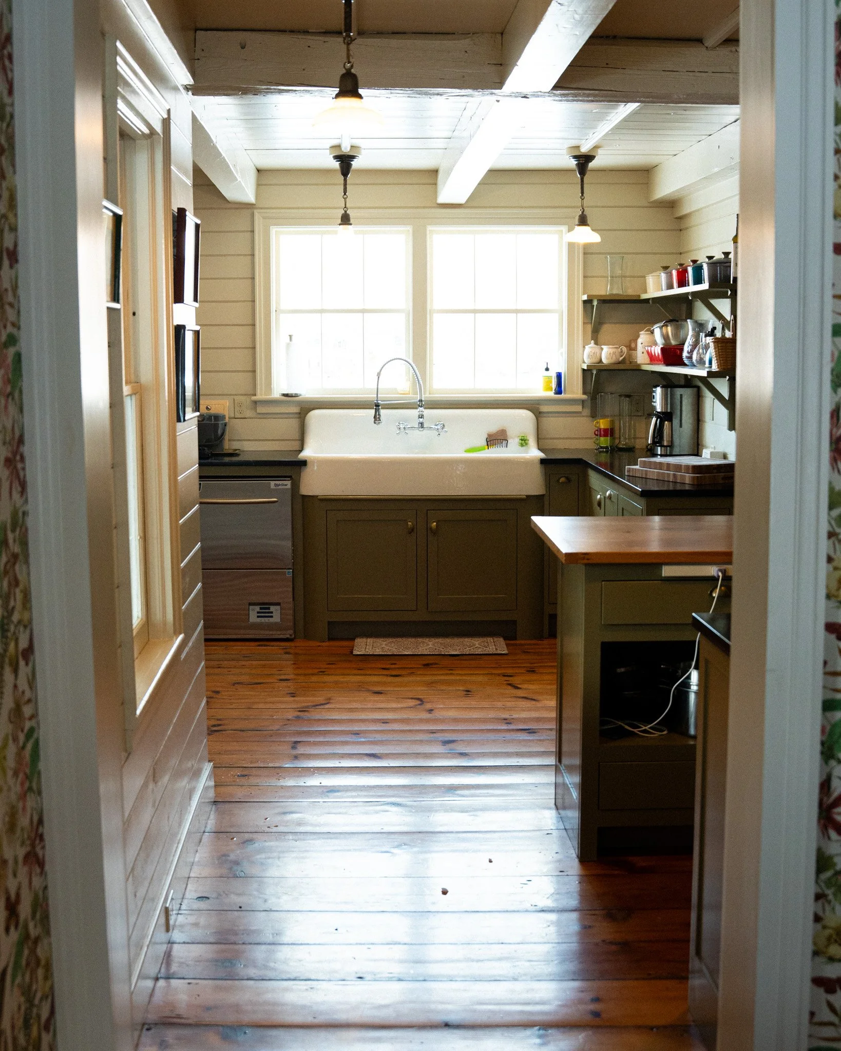 A cozy kitchen with wooden floors and white paneled walls, featuring a farmhouse sink beneath two windows, open shelving with dishes, a coffee maker, and a small kitchen island.