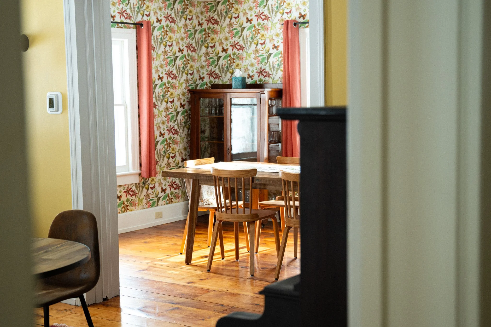 View into a dining room with floral wallpaper, a wooden dining table with chairs, a wooden cabinet with glass doors, and windows with coral curtains.