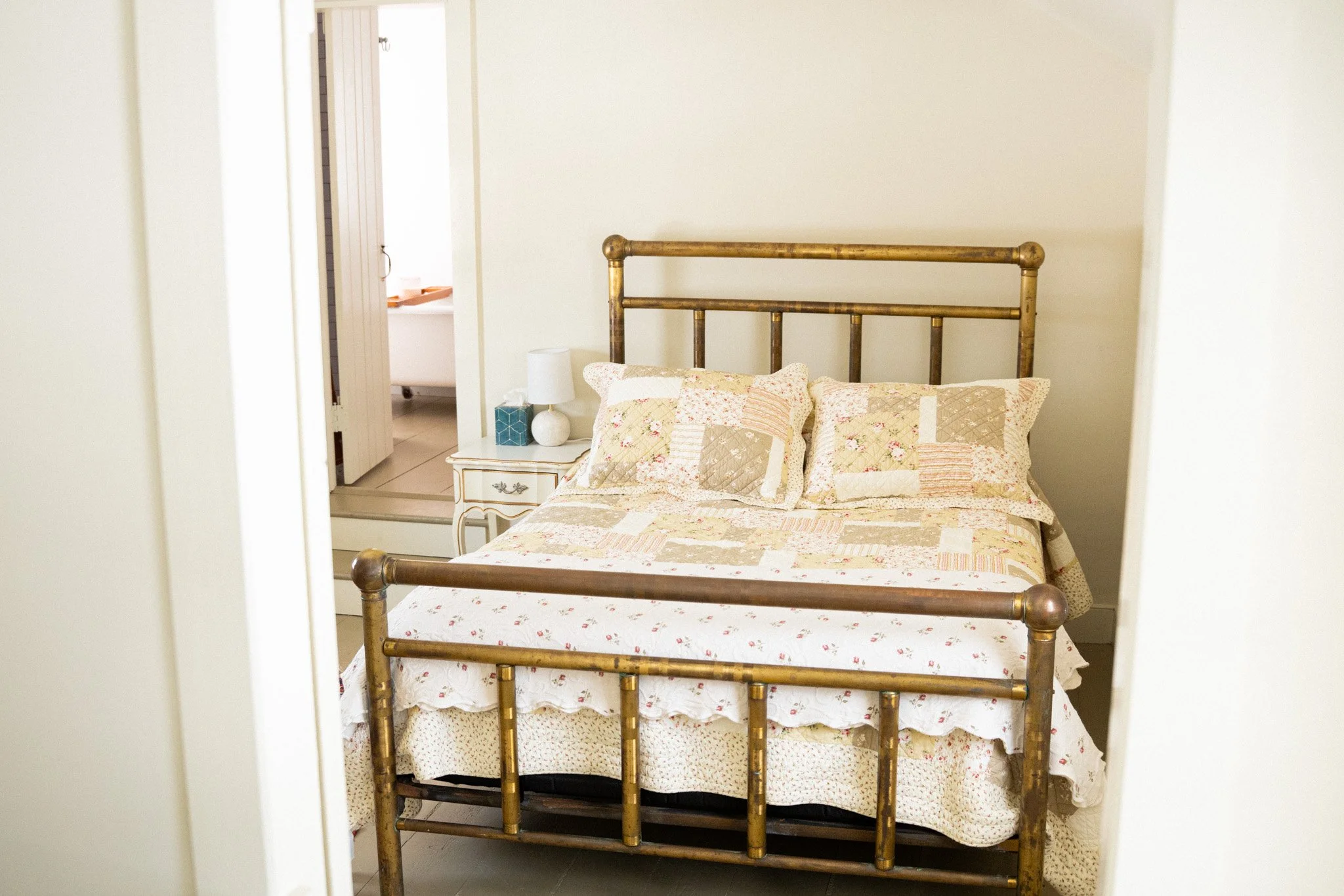 A bedroom with a brass bed, floral quilt, and matching pillows. A white nightstand with a lamp and tissue box is beside the bed. An open door reveals a bathroom with a bathtub.