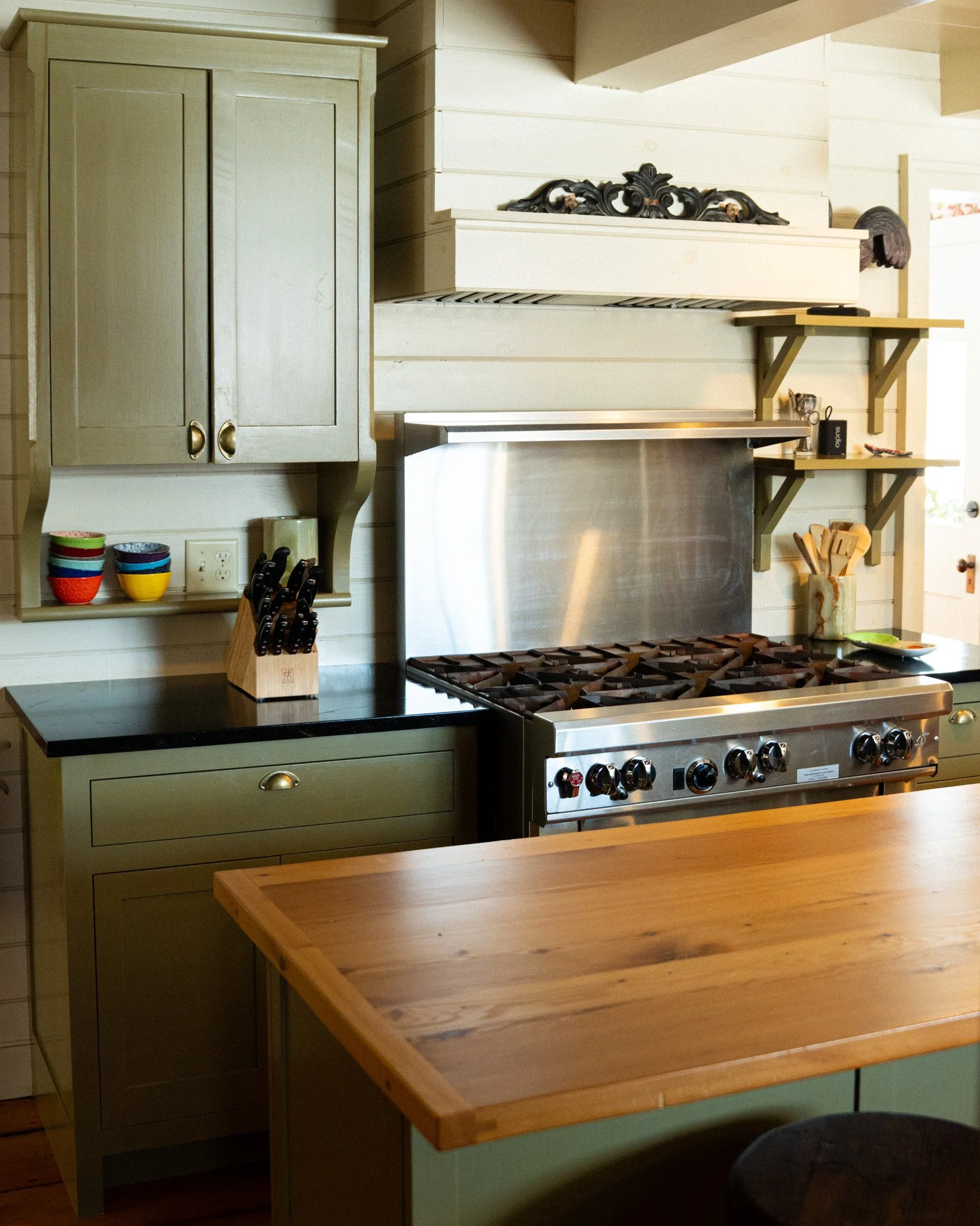 A kitchen with a gas stove, wooden countertops, open shelves, a green cabinet, and colorful bowls.