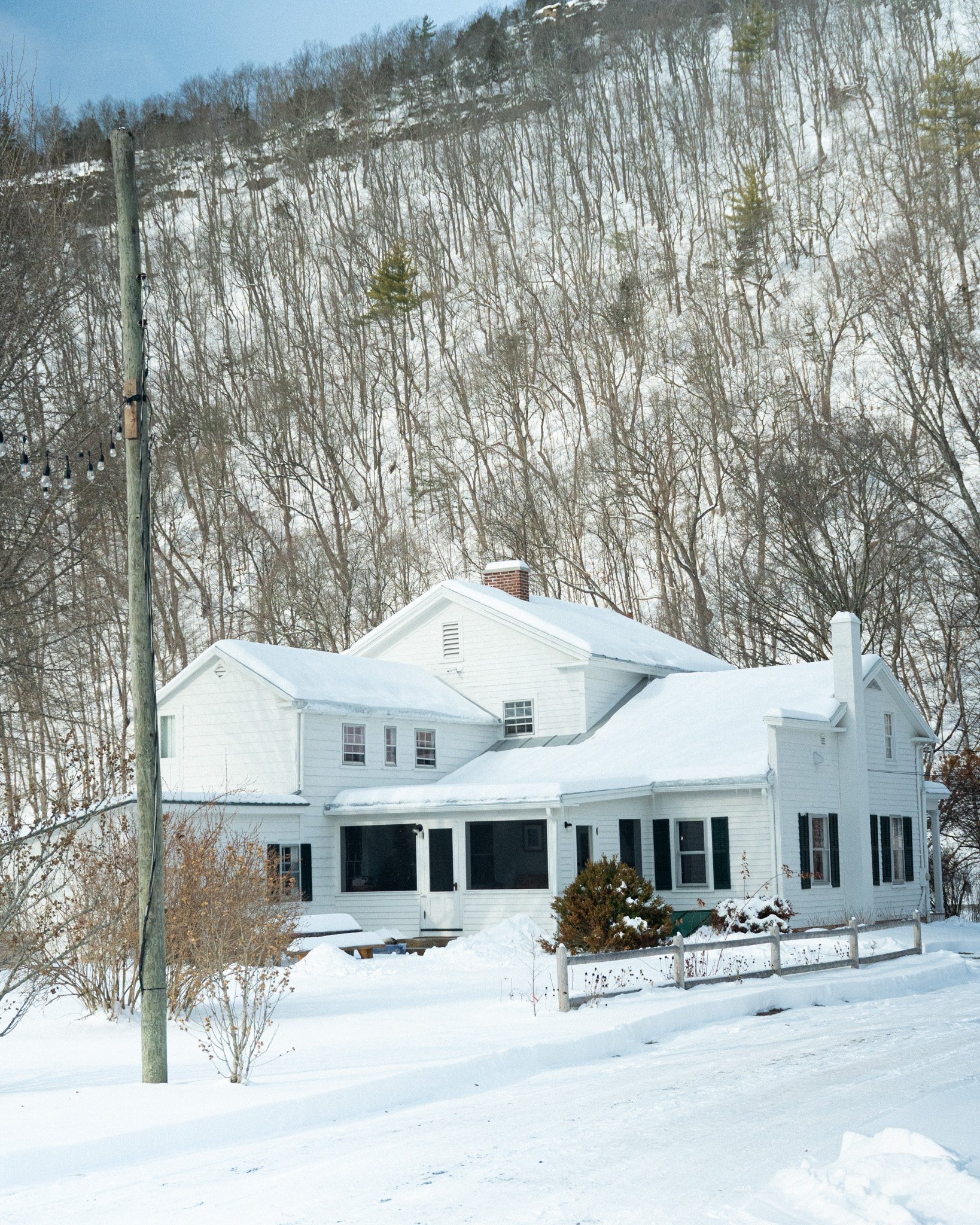 White house with snow-covered roof in a winter landscape, with a wooded hillside in the background, a utility pole with string lights, and a snow-covered yard with a small shrub and a wooden fence.