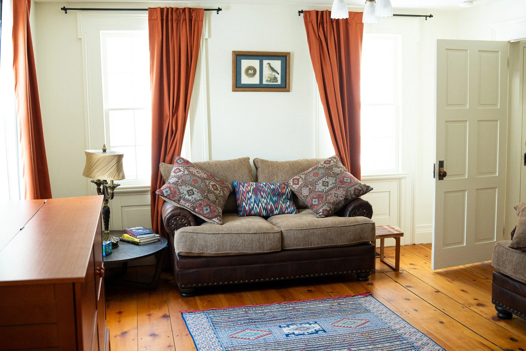 Living room with a beige sofa, colorful pillows, red curtains, wooden flooring, a side table with a lamp and books, and framed pictures on the wall.