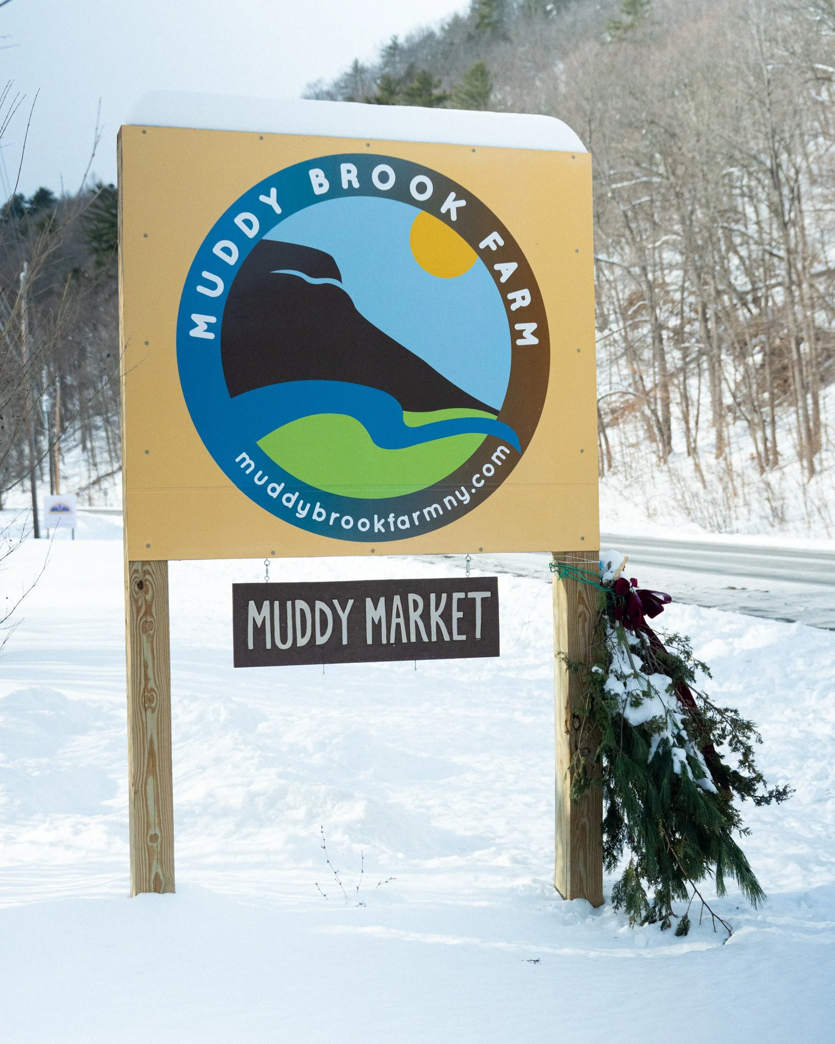 Sign for Muddy Brook Farm with a logo depicting a landscape including a hill, river, and sun, and text advertising the farm's website. Below the sign, a smaller sign reads "Muddy Market," decorated with greenery and snow.