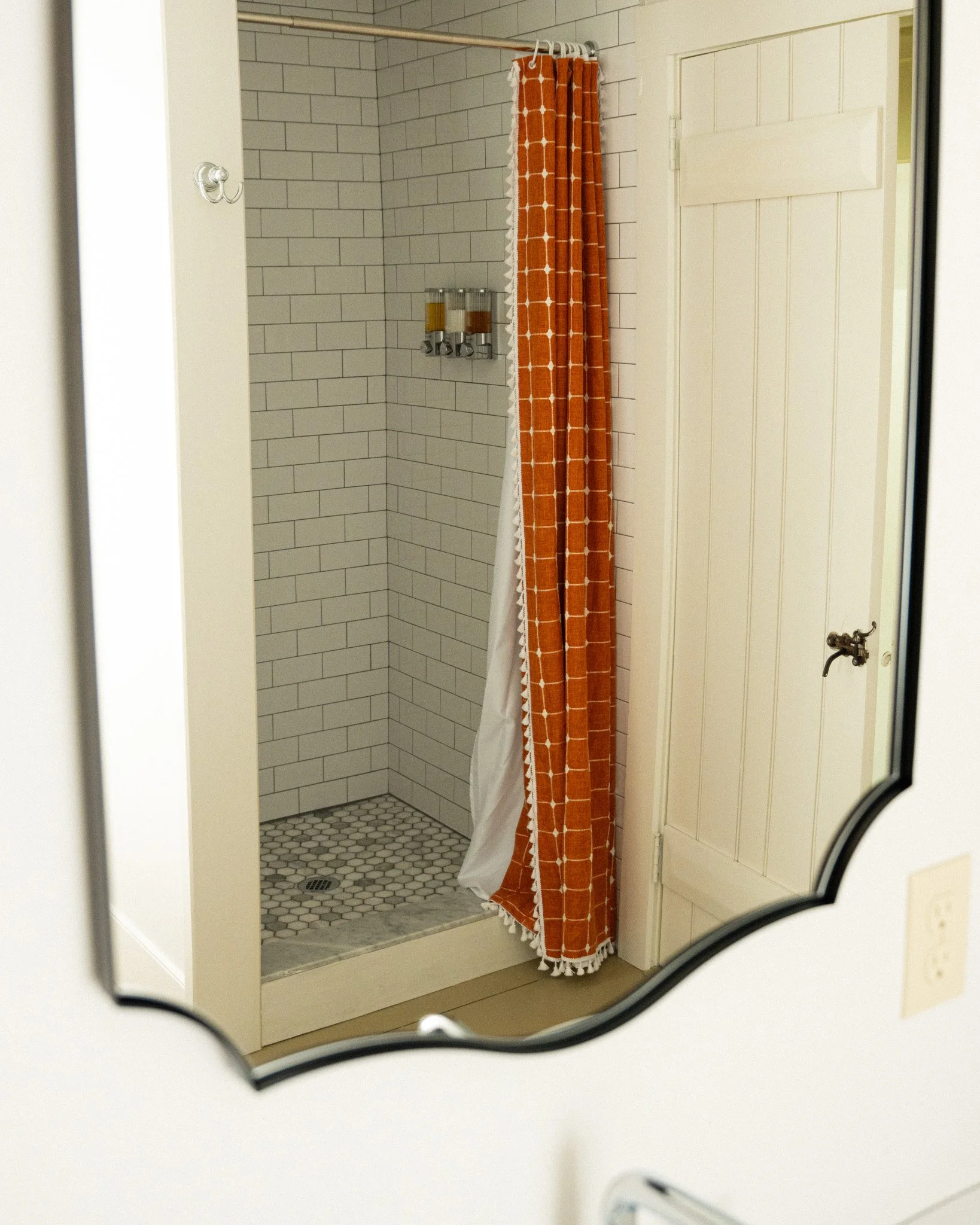 A small shower with white subway tiles, an orange patterned shower curtain, and dual dispensers on the wall inside.